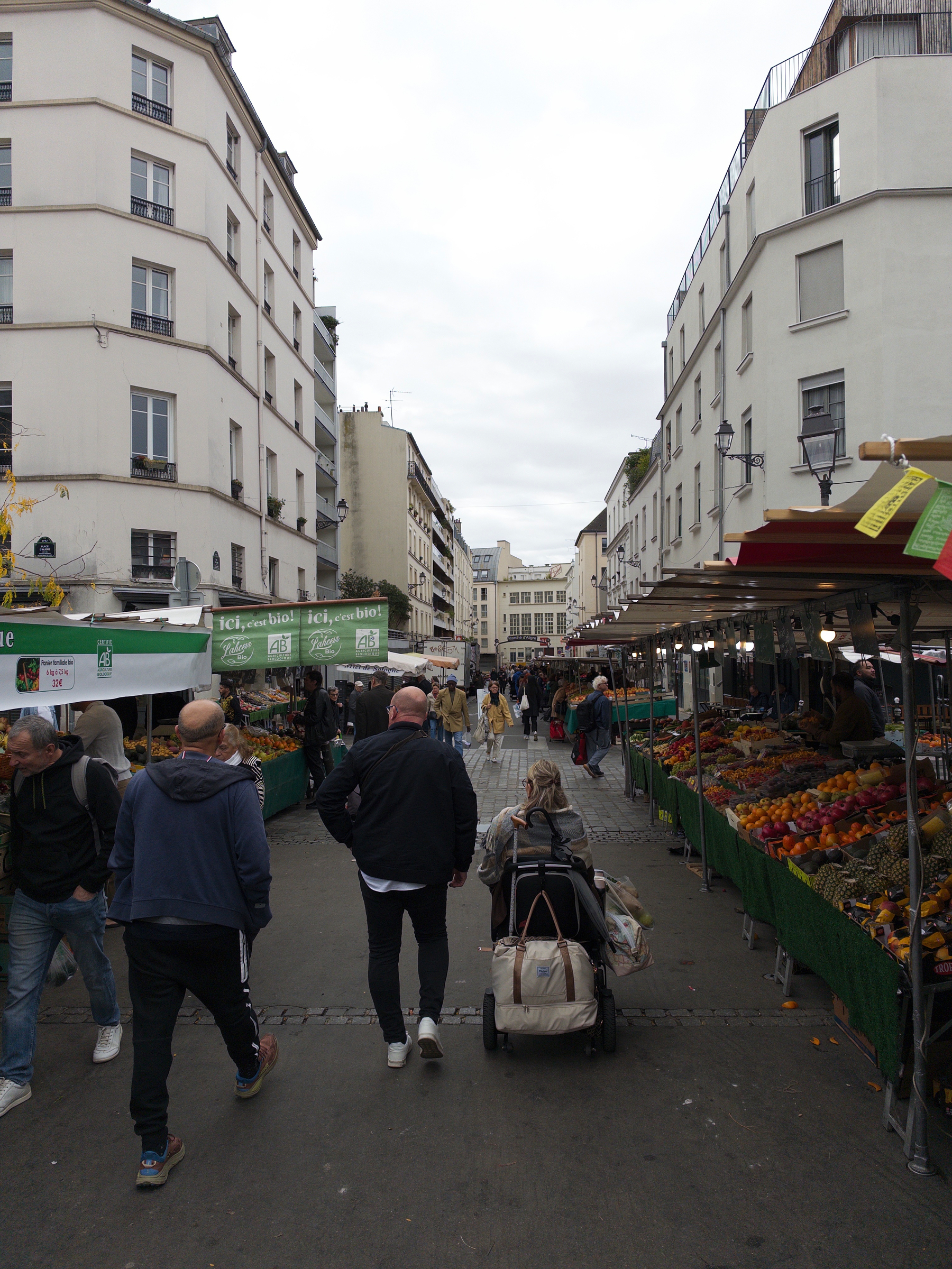 A bustling outdoor market street is lined with stalls selling fruits and vegetables, while people walk and shop amidst tall residential buildings.
