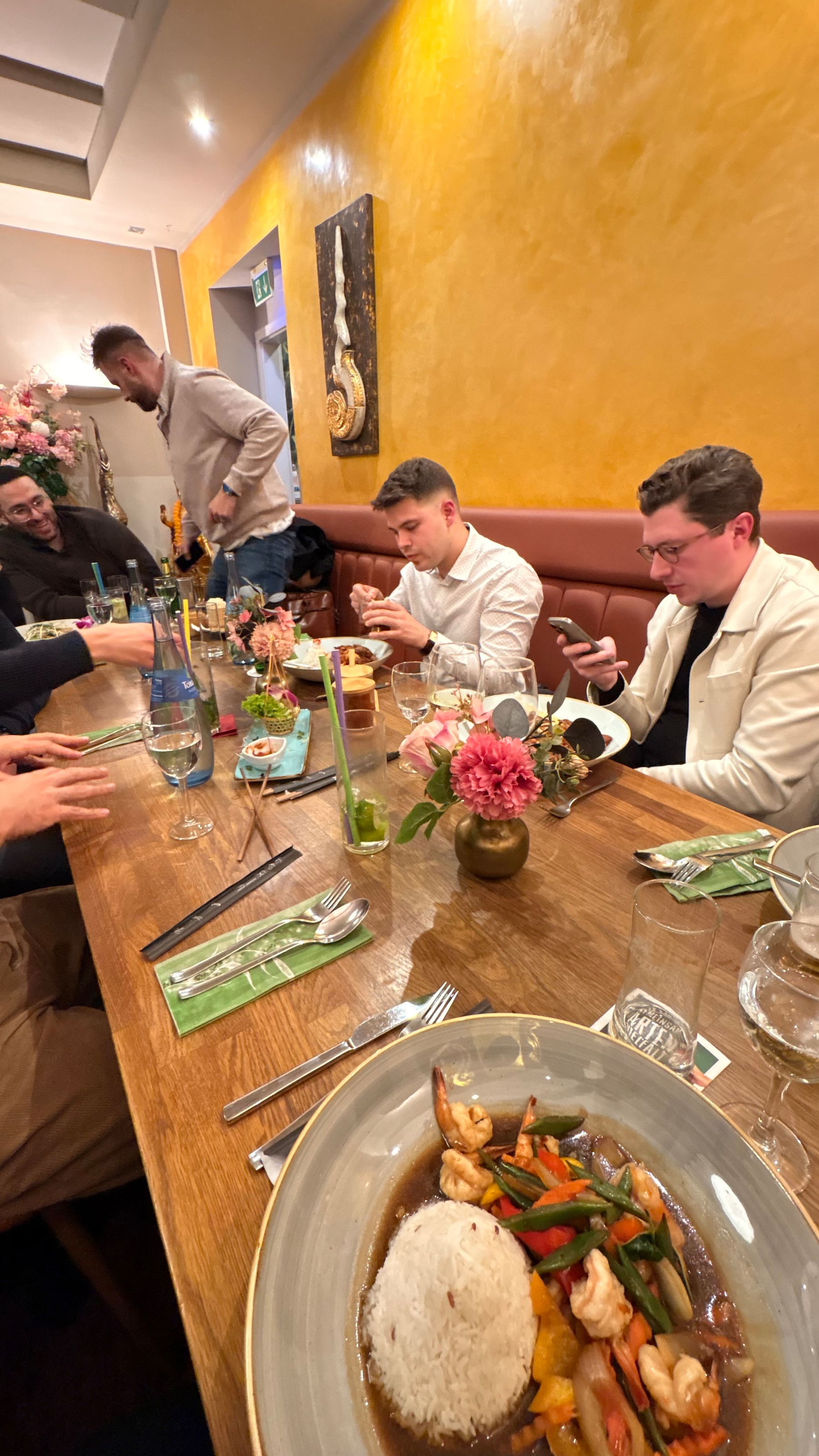 People are seated at a restaurant table with dishes of food, drinks, and floral decorations.