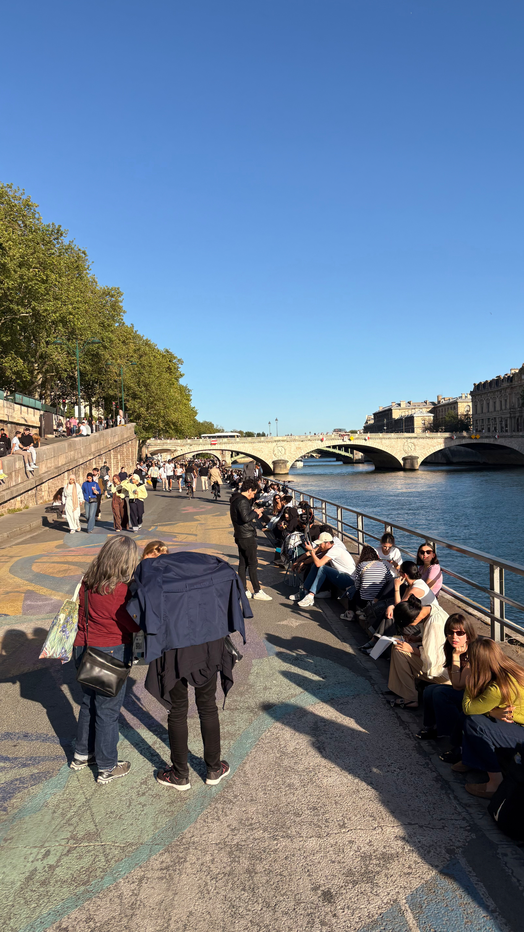 People are gathered along a riverside walkway under a clear blue sky, with a bridge and trees in the background.