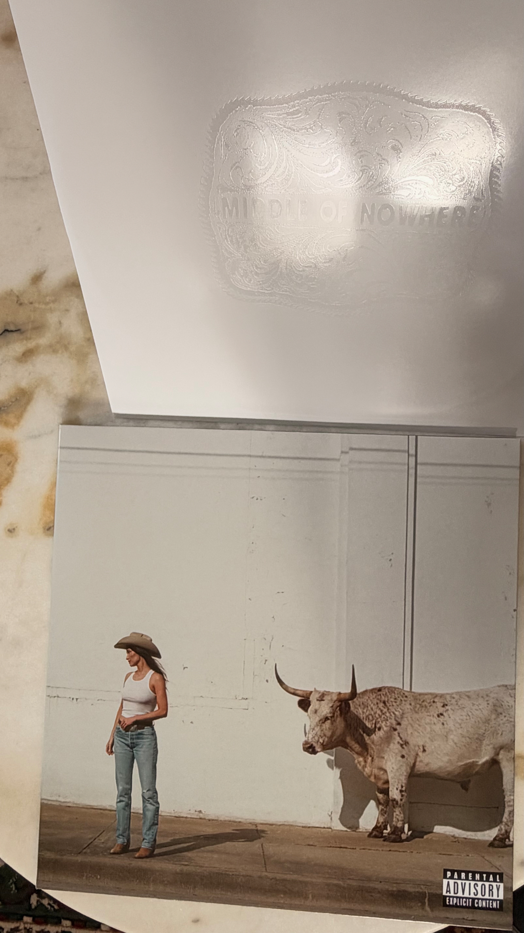 Auto-generated description: A woman wearing a cowboy hat, white tank top, and jeans stands on a sidewalk next to a large longhorn steer against a plain white wall.