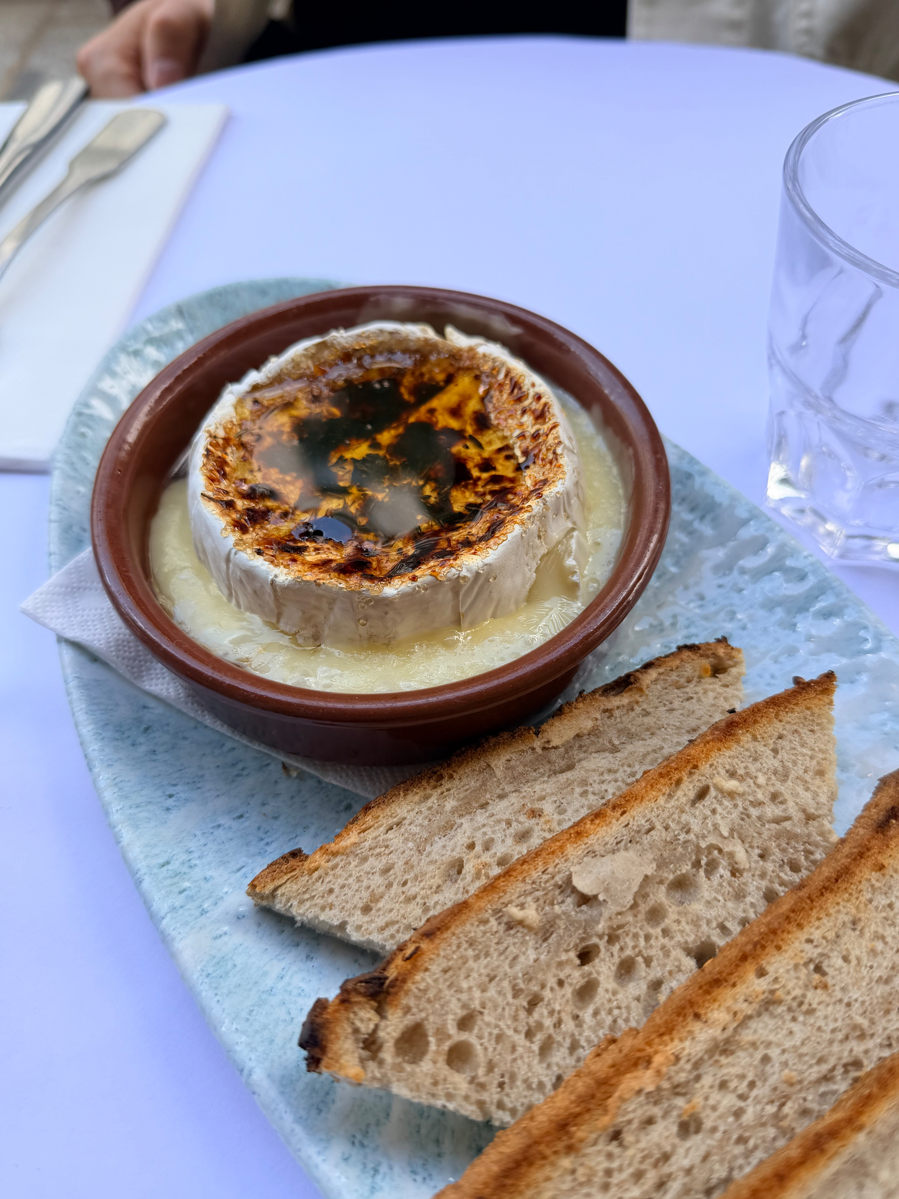 Auto-generated description: A serving of baked cheese with a caramelized top is paired with slices of grilled bread on a plate beside a glass and cutlery.
