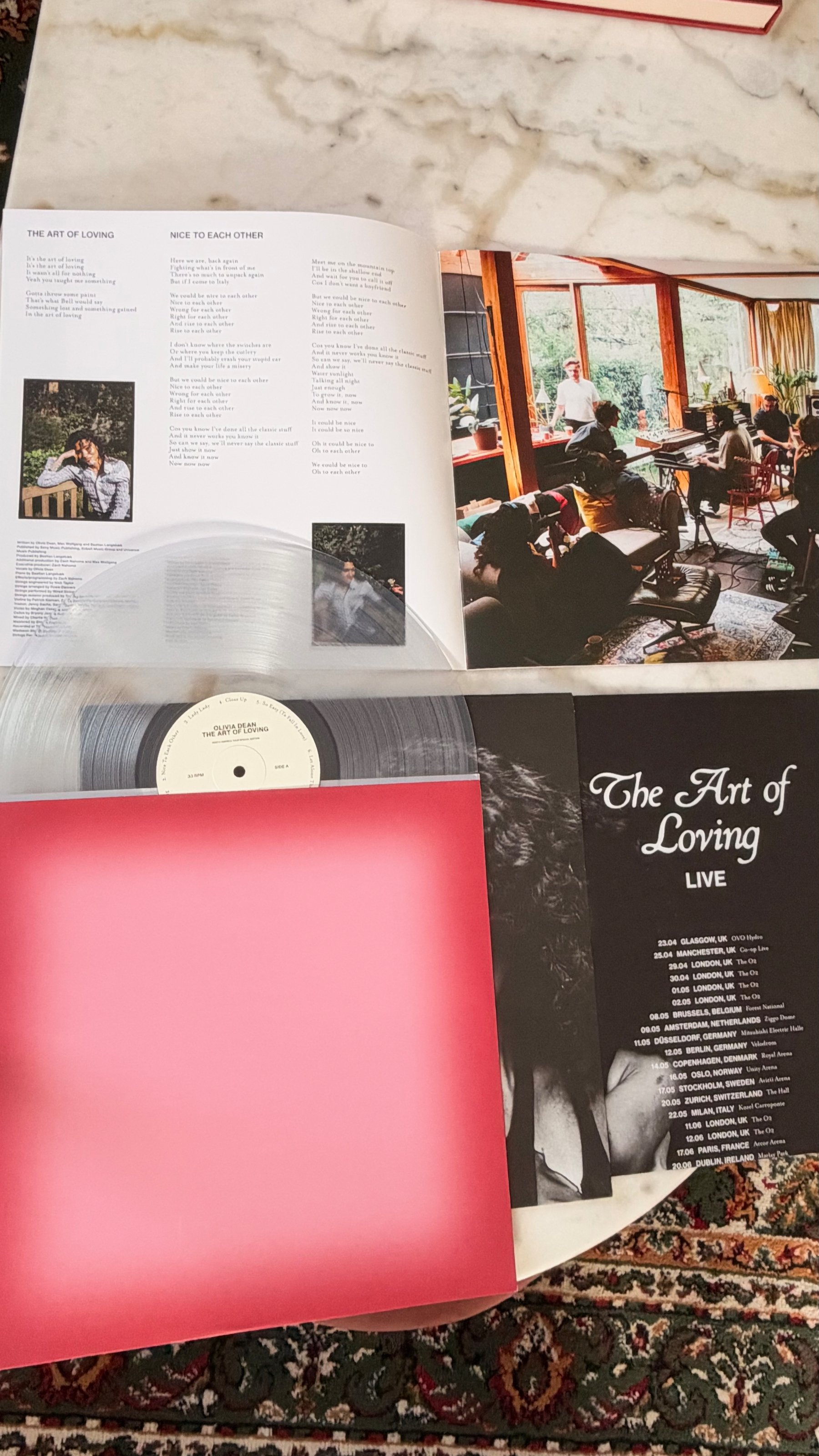 Auto-generated description: A clear vinyl record and its album cover are displayed on a marble surface alongside an open booklet with song lyrics and a black and white flyer titled The Art of Loving Live showing concert dates.
