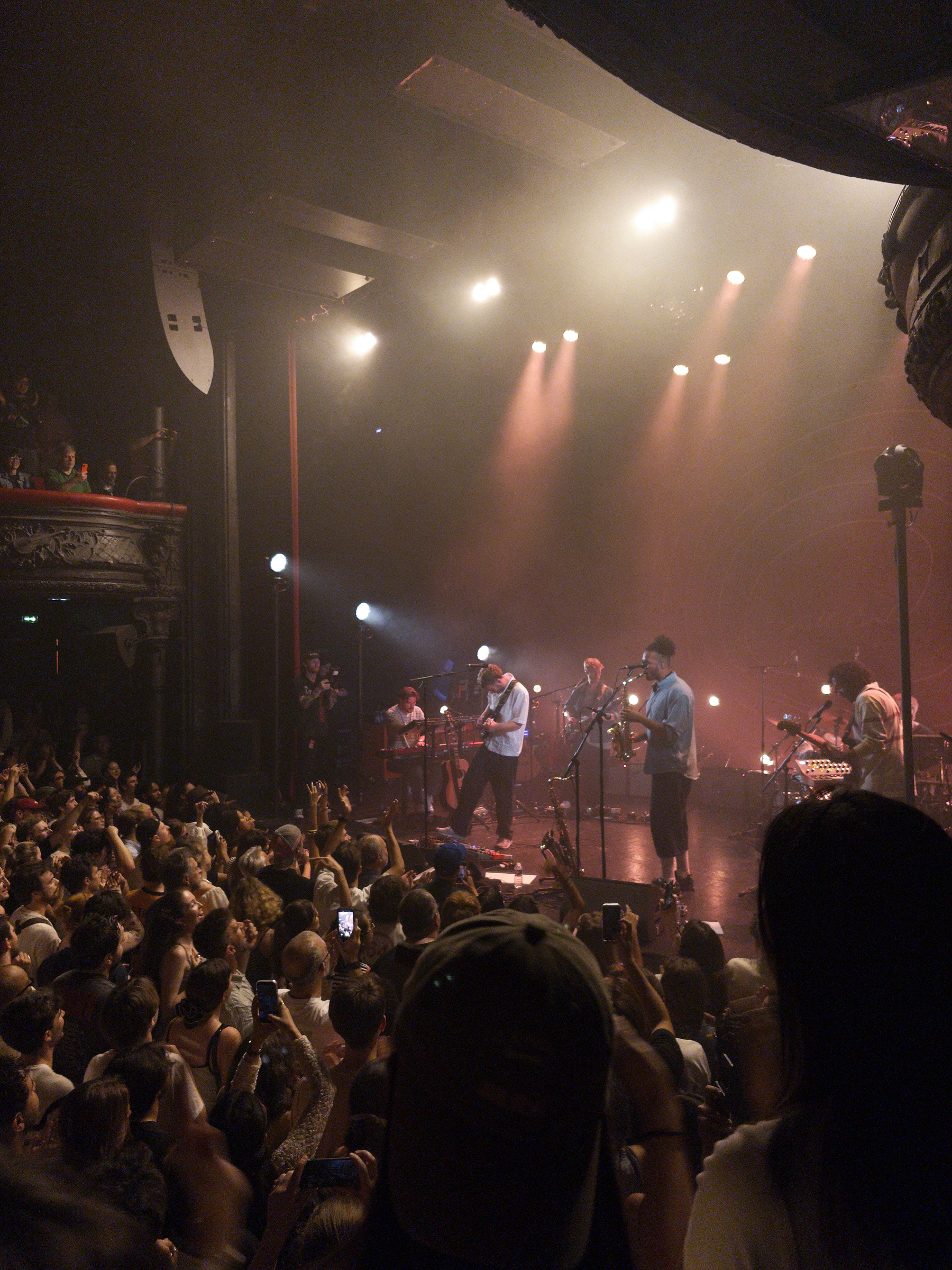Auto-generated description: A live band performs on a dimly lit stage surrounded by an enthusiastic audience in a crowded concert venue.