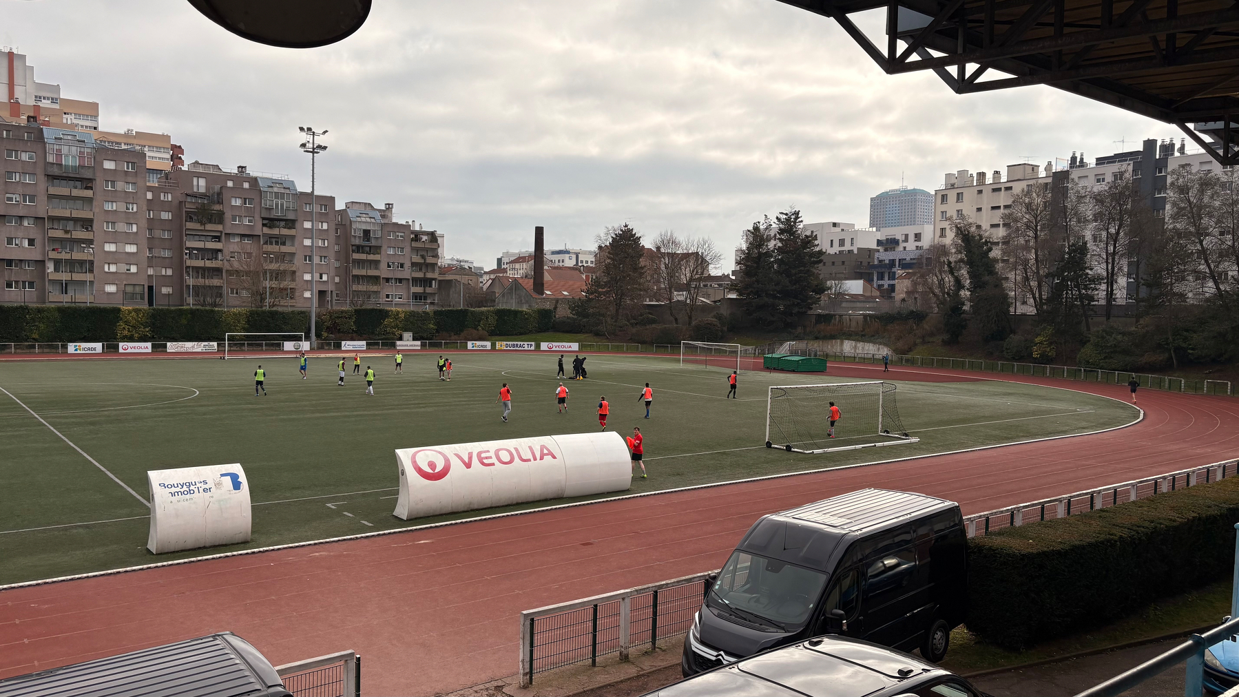 A soccer field surrounded by a running track hosts a soccer match with players in brightly colored jerseys, set against an urban backdrop with buildings and overcast skies.