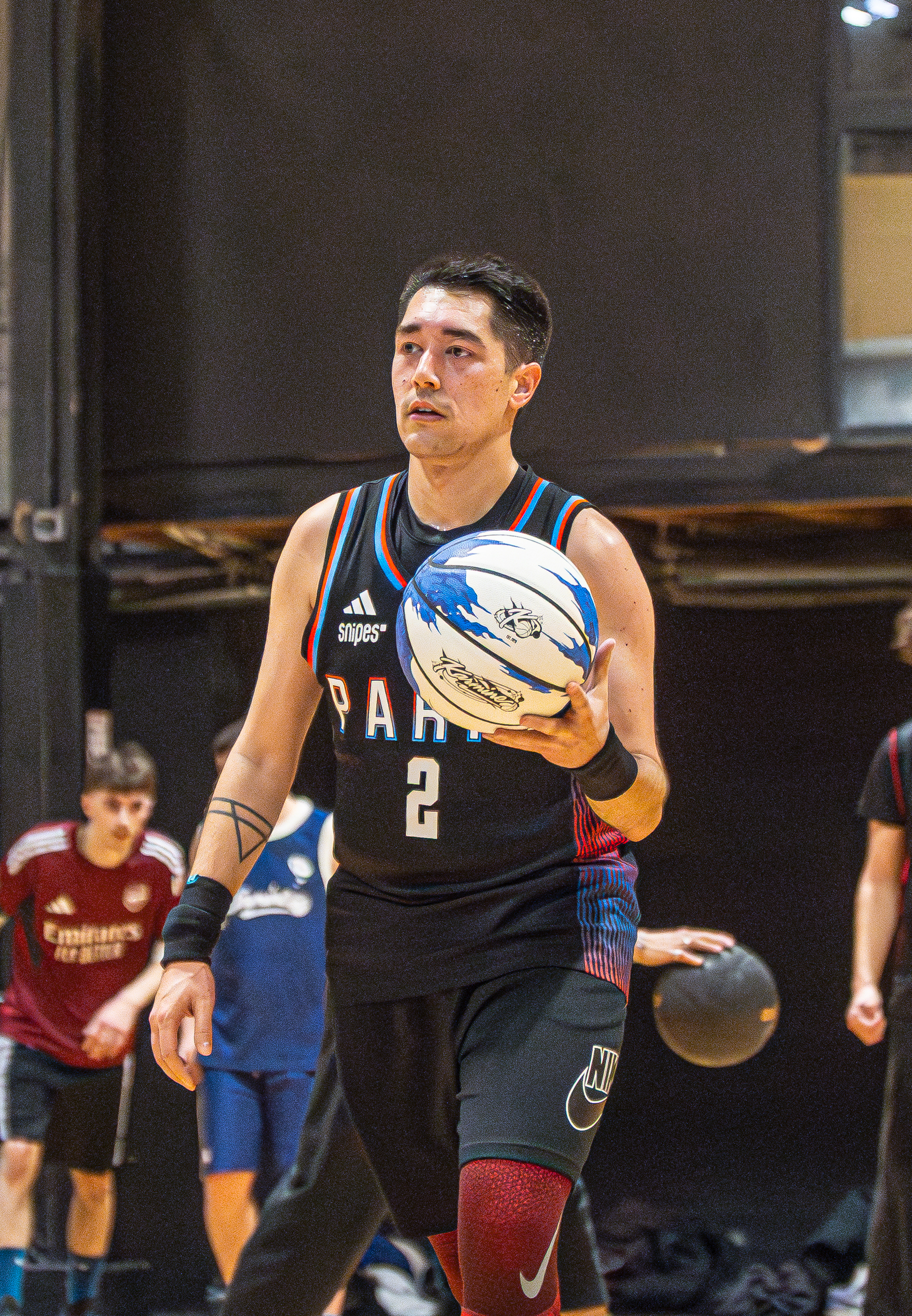 A basketball player wearing a black jersey dribbles a blue and white ball on a court, with other players in the background.