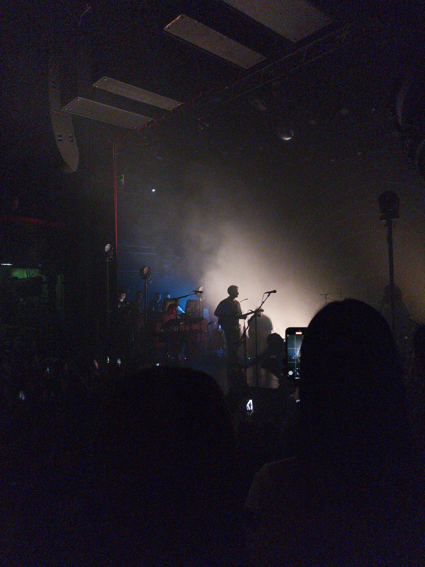 Auto-generated description: A band performs on a dimly lit stage surrounded by a foggy atmosphere with an audience capturing the moment.