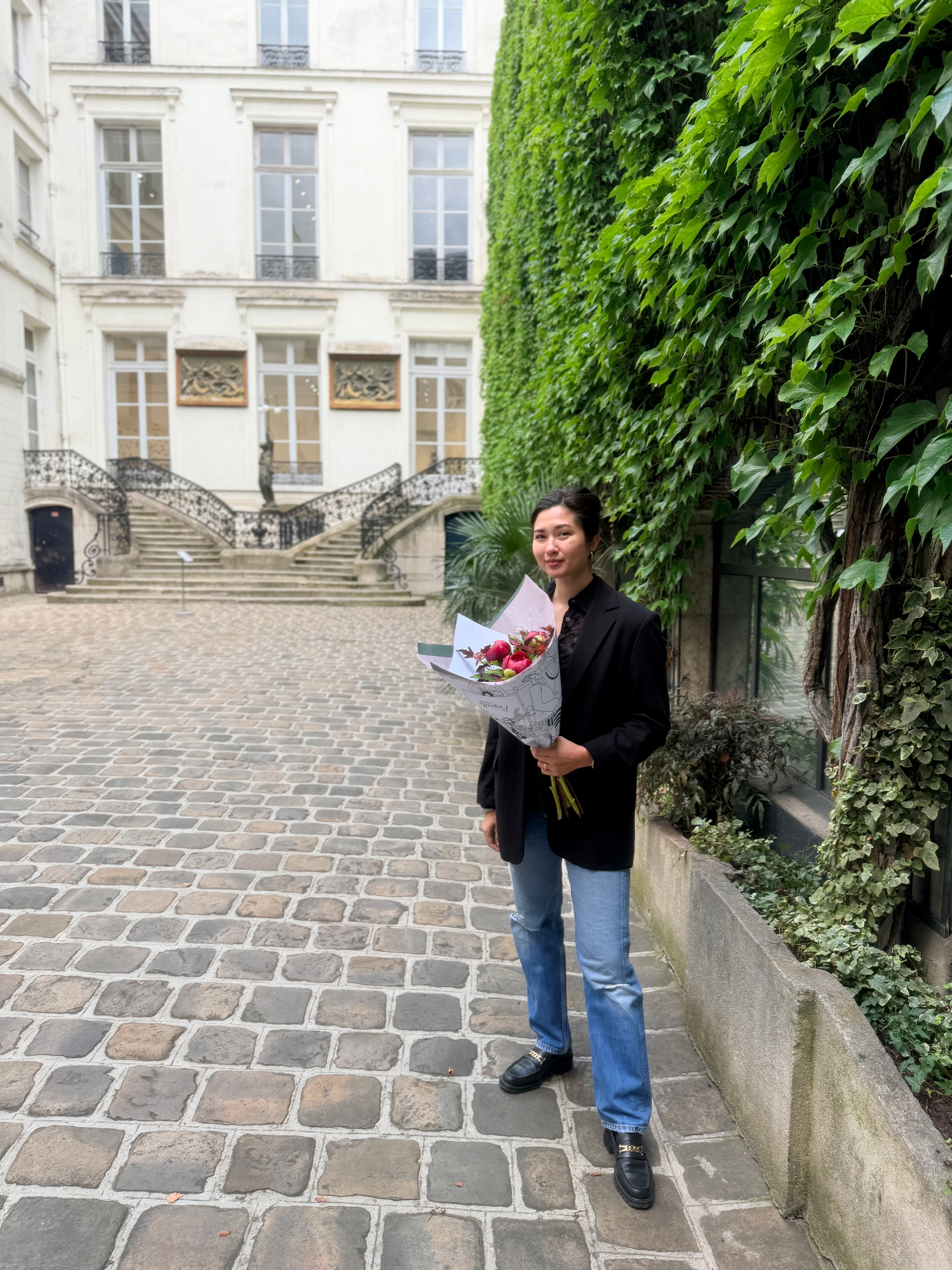 Auto-generated description: A person holding a bouquet of flowers stands on a cobblestone path in front of a building with ivy-covered walls.