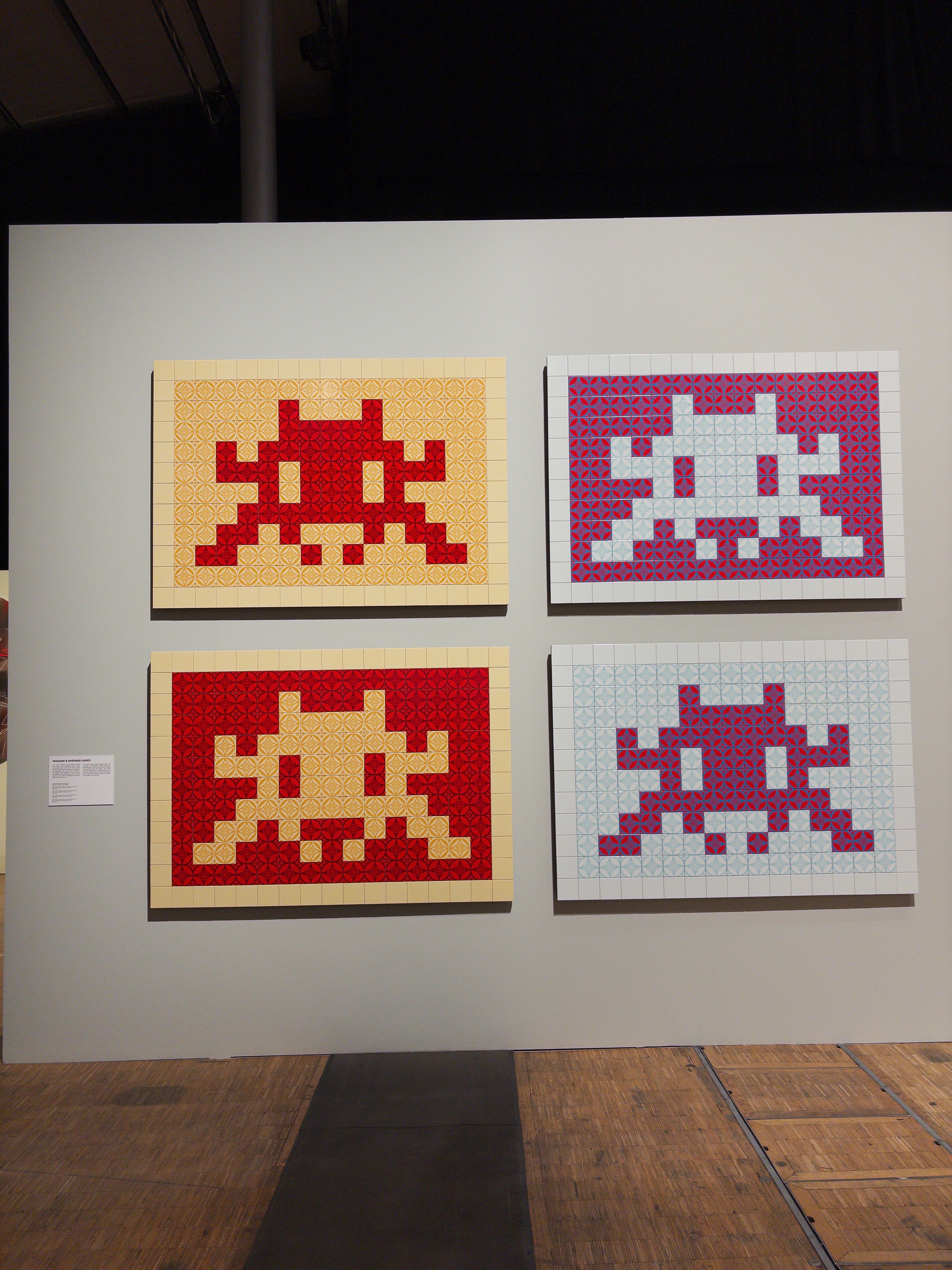 Four pixelated alien figures are displayed on a wall, each created with contrasting colored squares on individual rectangular canvases.
