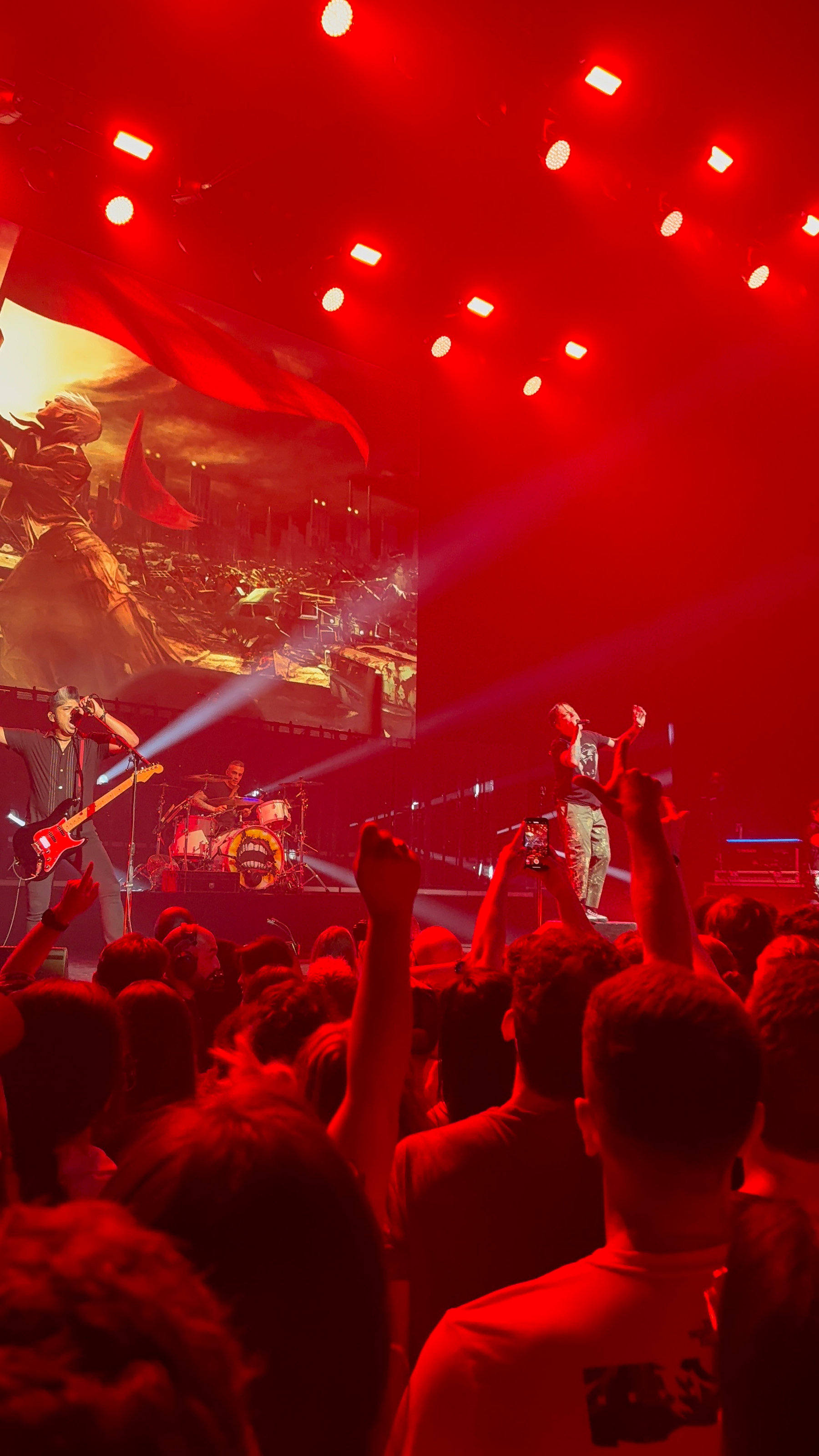 Auto-generated description: A live concert is underway with a band performing on stage under red lighting while an enthusiastic crowd watches and records the event.