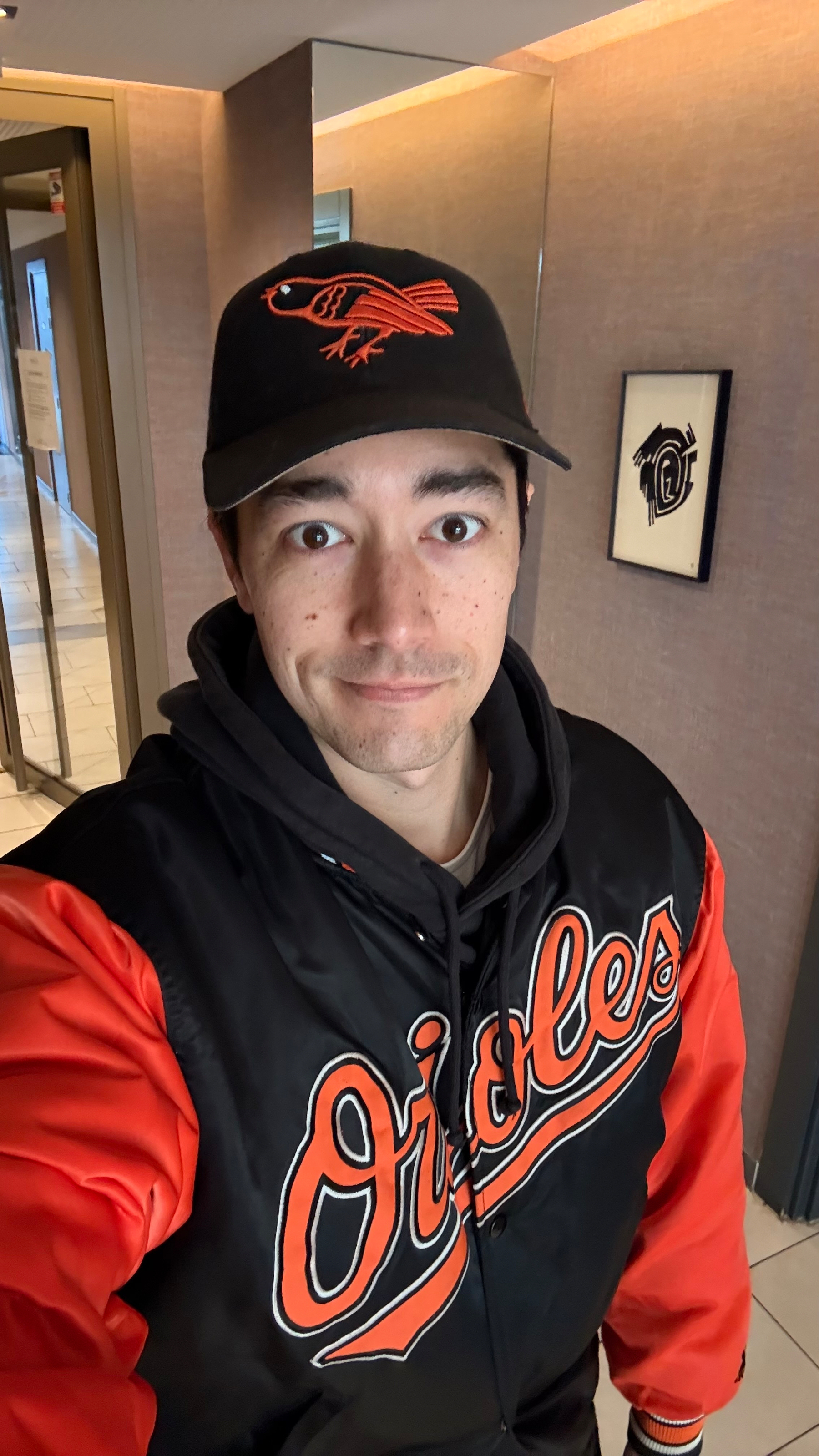 A person wearing an Orioles jacket and cap is standing indoors, smiling at the camera.