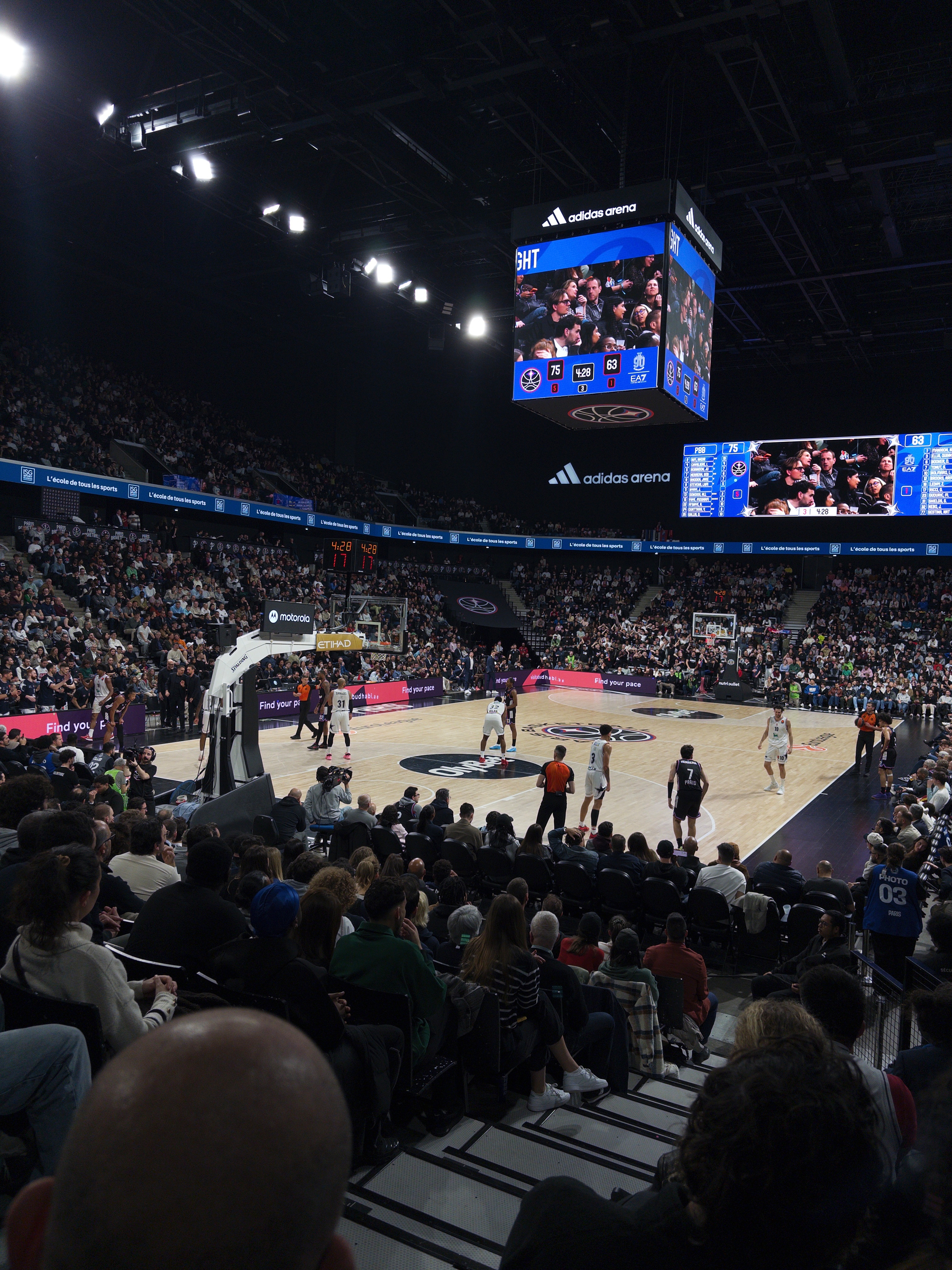 A basketball game is taking place in a large indoor arena filled with spectators, featuring teams in contrasting uniforms and a prominently displayed scoreboard.