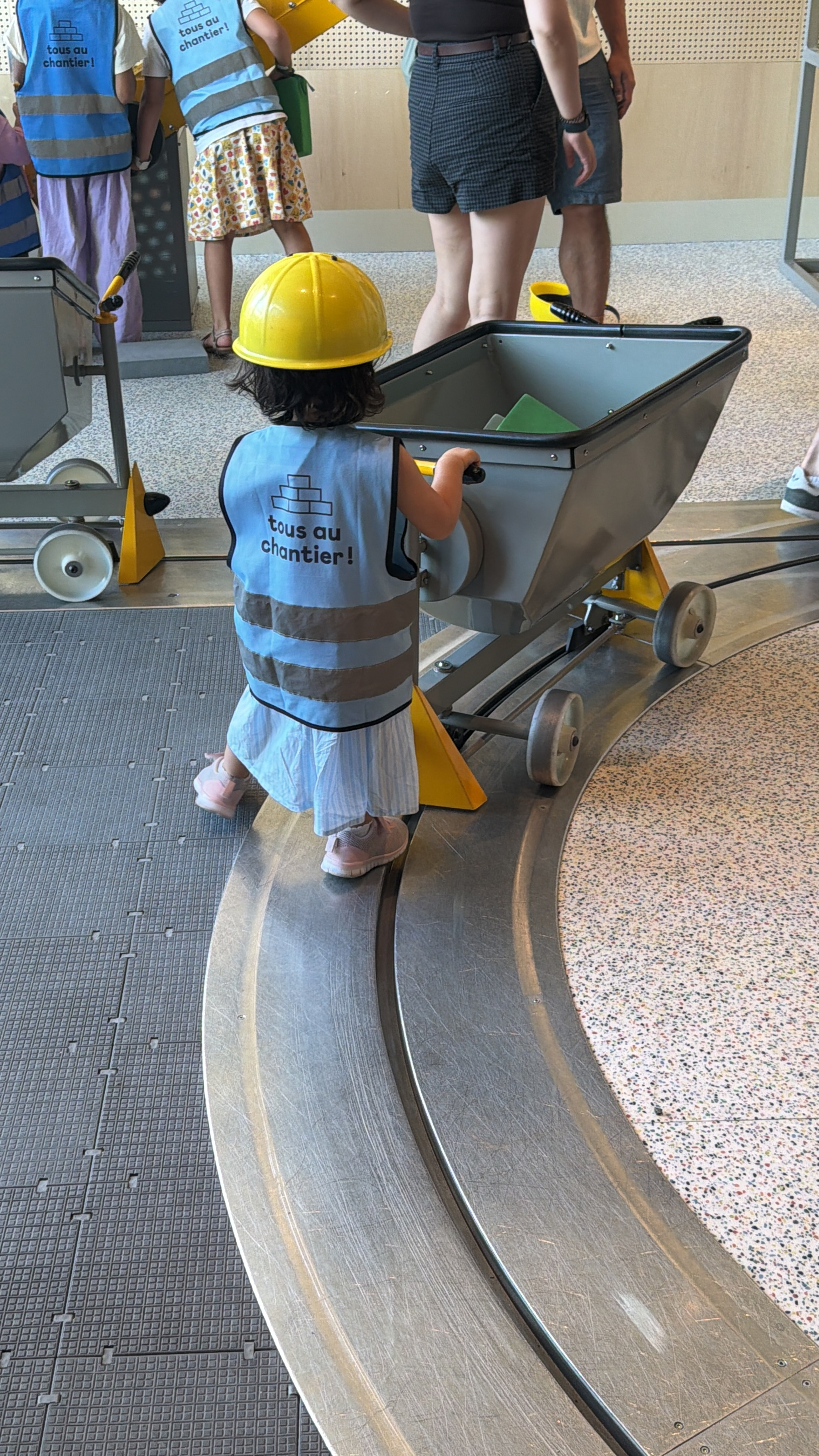 Auto-generated description: A young child wearing a yellow construction helmet and a blue vest labeled tous au chantier! is pushing a small gray cart along a curved track, surrounded by other children and adults.