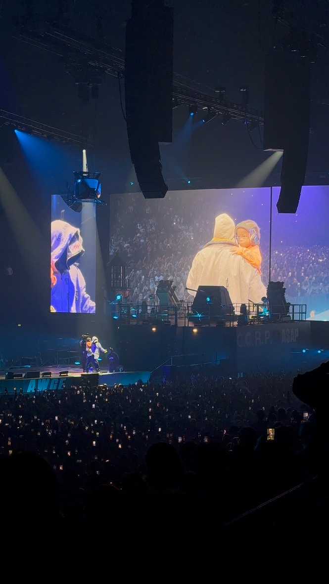 A large crowd watches a performer holding a child on stage at a concert, with the scene magnified on big screens above.