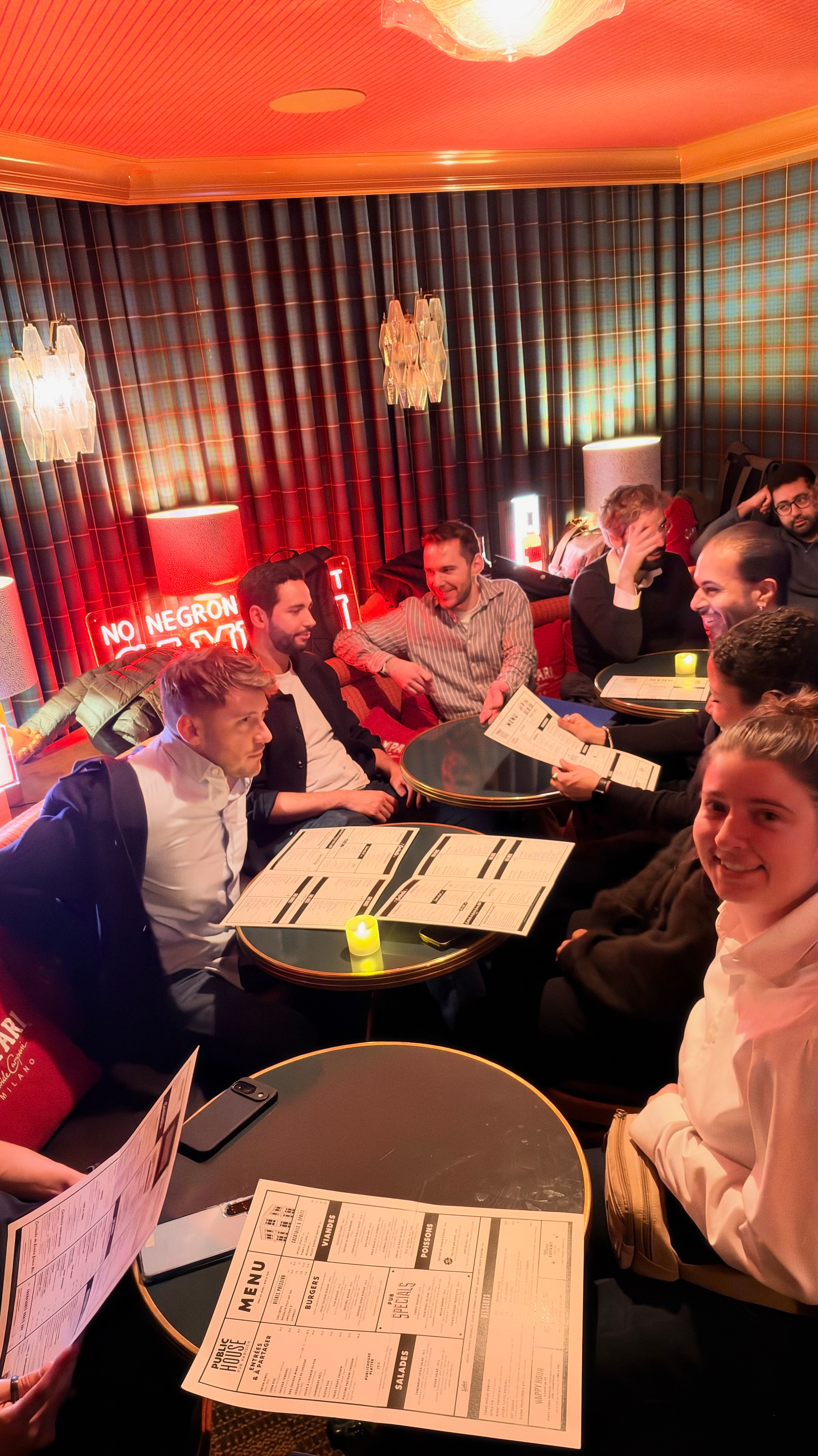 A group of people is gathered around tables in a cozy, warmly lit room, looking at menus and engaging in conversation.