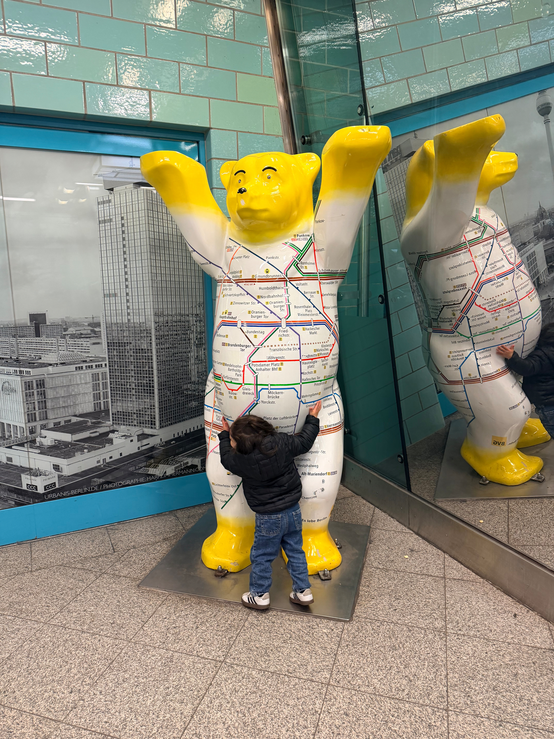 Auto-generated description: A child is hugging a large yellow bear statue covered in a map design inside a modern-looking building.