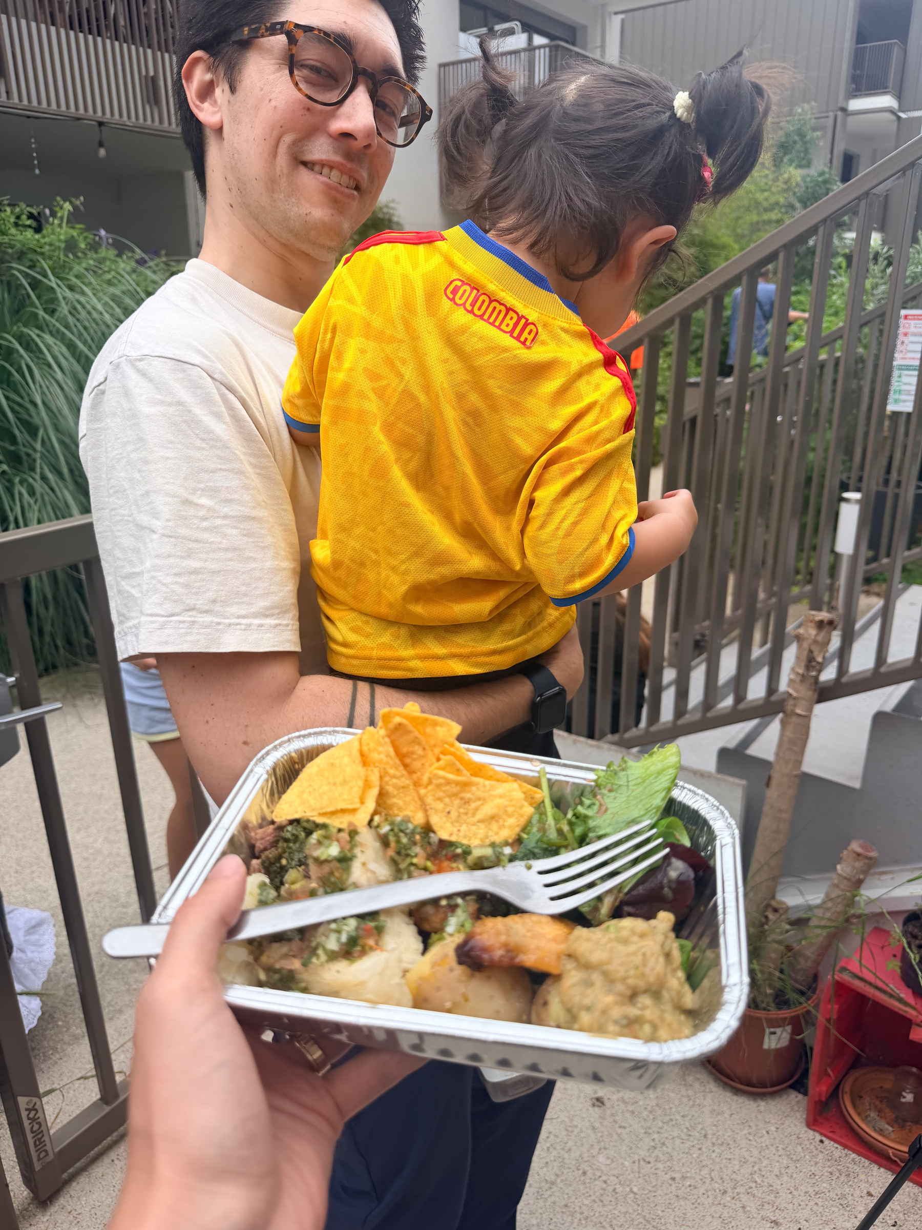 A person holding a child wearing a yellow jersey, while another hand holds a tray of mixed food, outdoors near a staircase and plants.