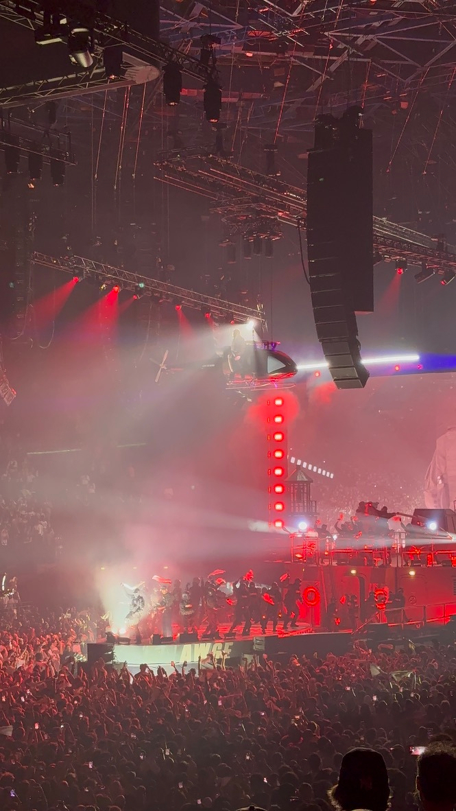 A large crowd watches a concert performance with intense red lighting and a person suspended above the stage by a helicopter-like device.