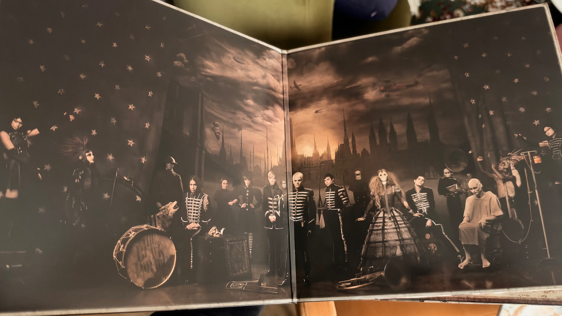 A group of people dressed in dark, theatrical costumes are posed in a moody, gothic setting with a cityscape background under a cloudy sky.