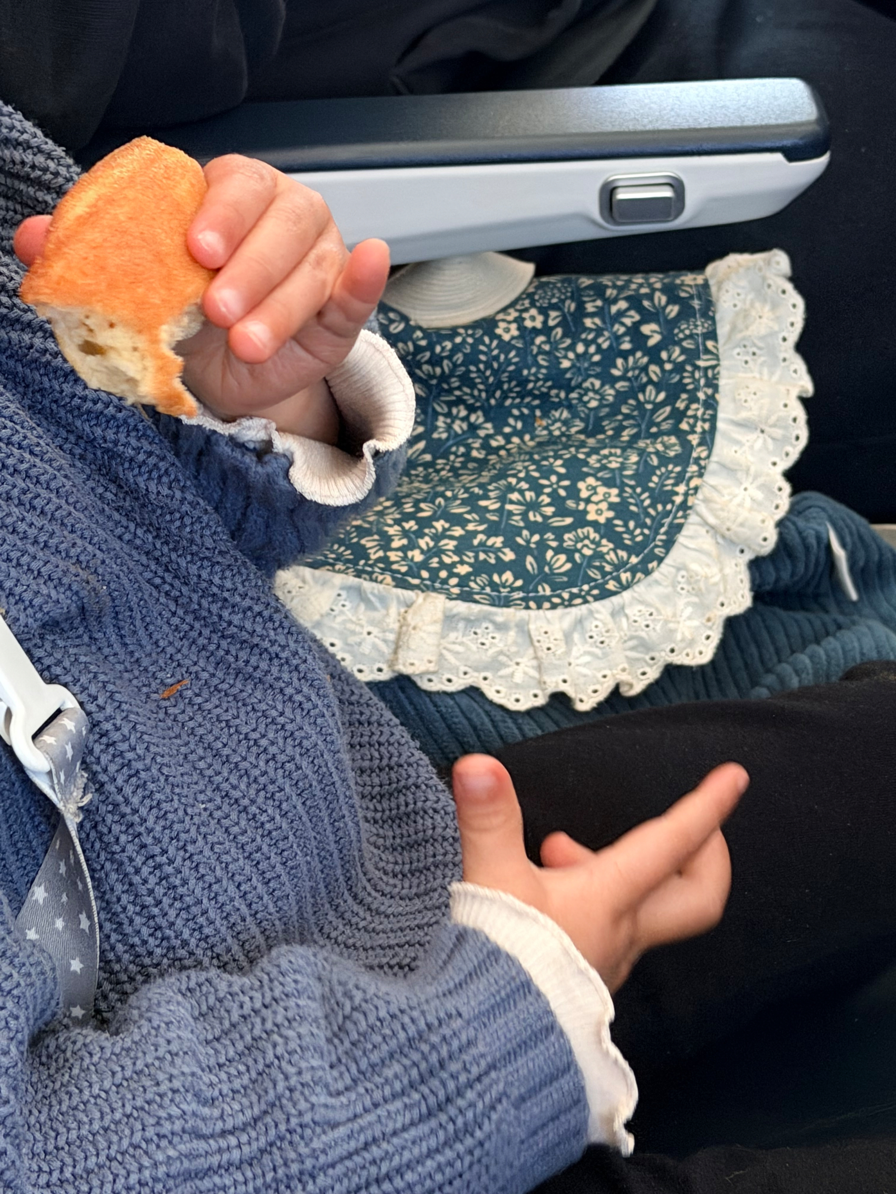 Auto-generated description: A child wearing a blue sweater is holding a piece of bread while seated on a lap with a blue and white patterned fabric underneath.