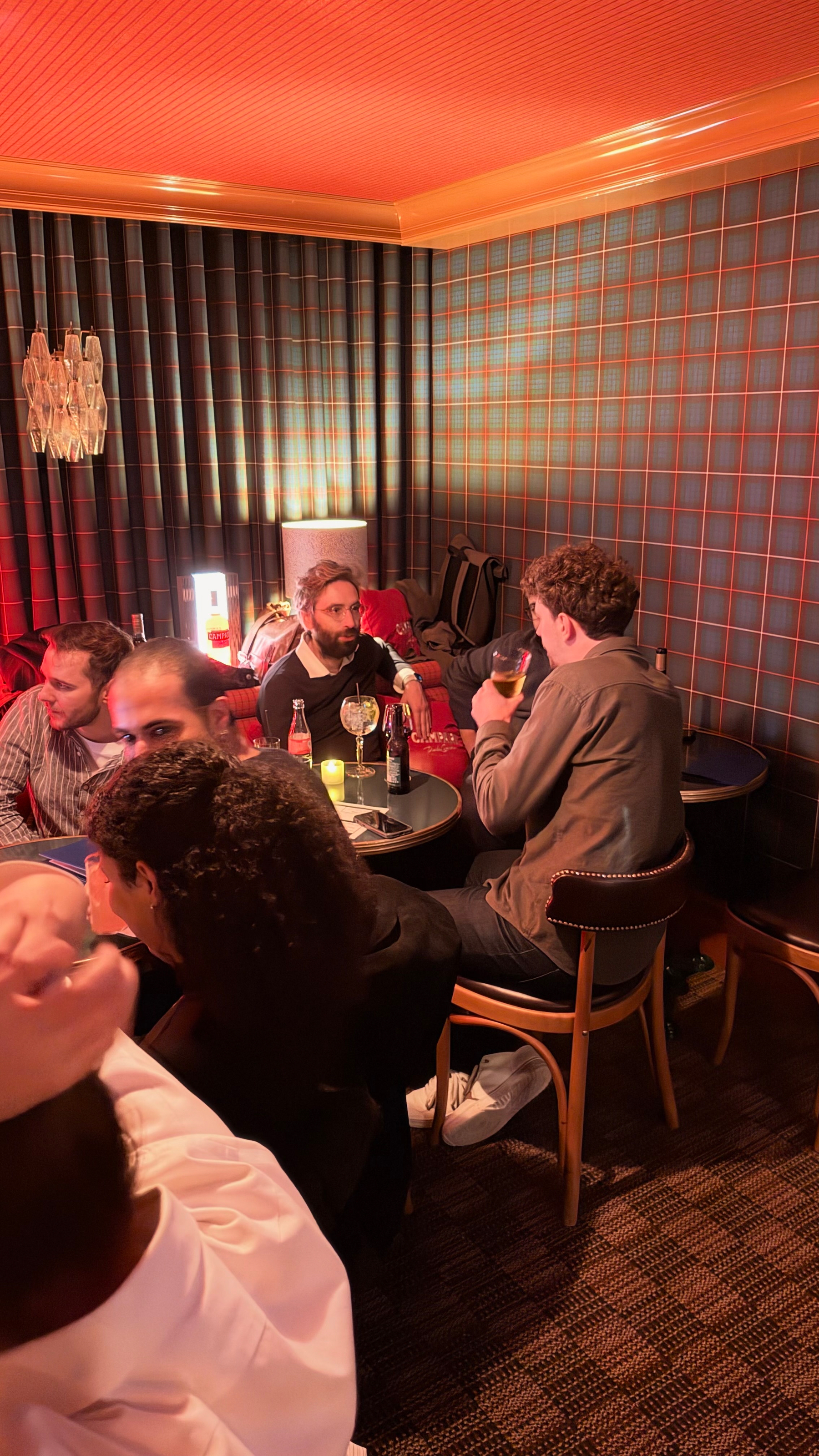 A group of people are socializing and enjoying drinks in a cozy, dimly lit room with patterned walls and warm lighting.