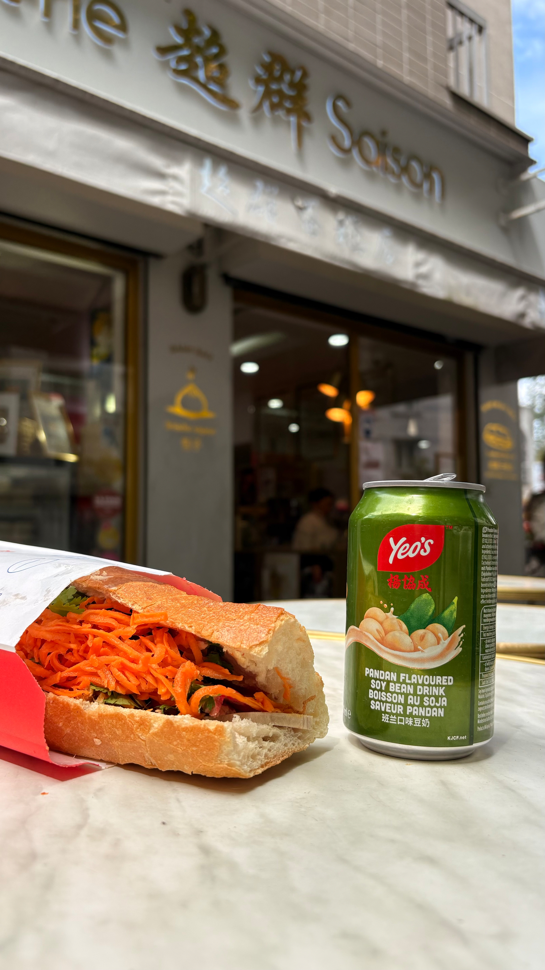 A sandwich filled with shredded carrots and greens is placed next to a can of Yeo's pandan-flavored soy bean drink on a marble table outside a shop.