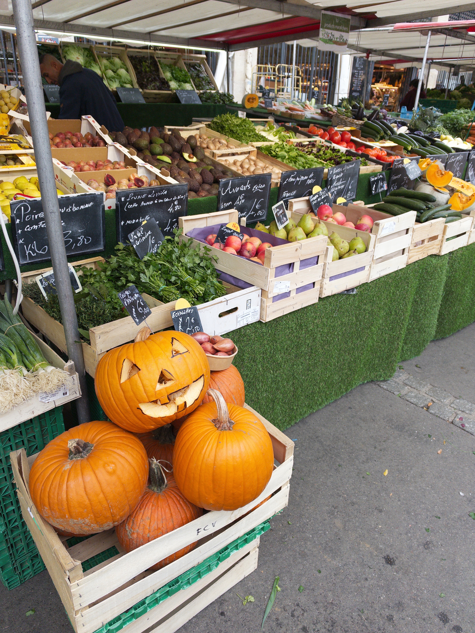 A market stall displays a variety of fresh fruits and vegetables, including a crate of pumpkins, one of which is carved into a jack-o'-lantern.