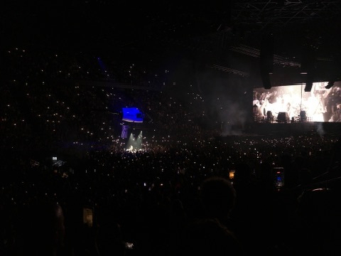 A large crowd is attending a concert with a brightly lit stage and a screen displaying visuals in a darkened arena.