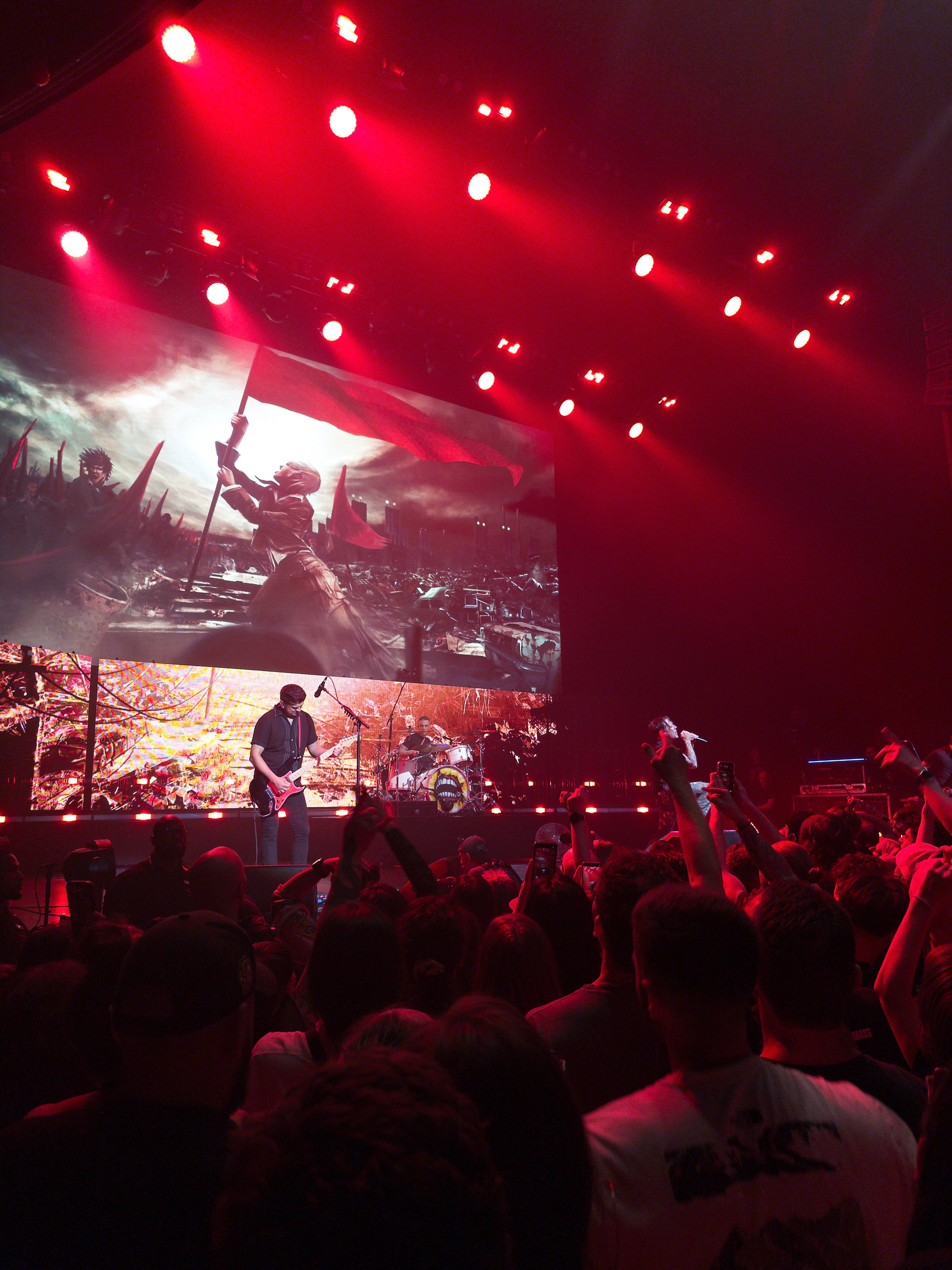 Auto-generated description: A band is performing on a stage bathed in red lights with a large screen behind them displaying dramatic imagery of a historical or battle scene.