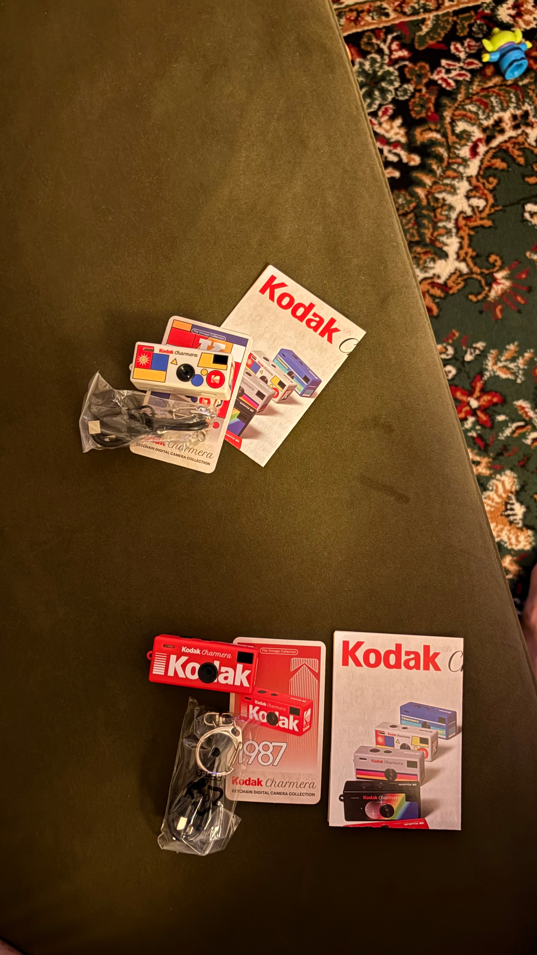 Two Kodak-themed digital camera keychains with accompanying packaging and manuals are placed on a green fabric surface.