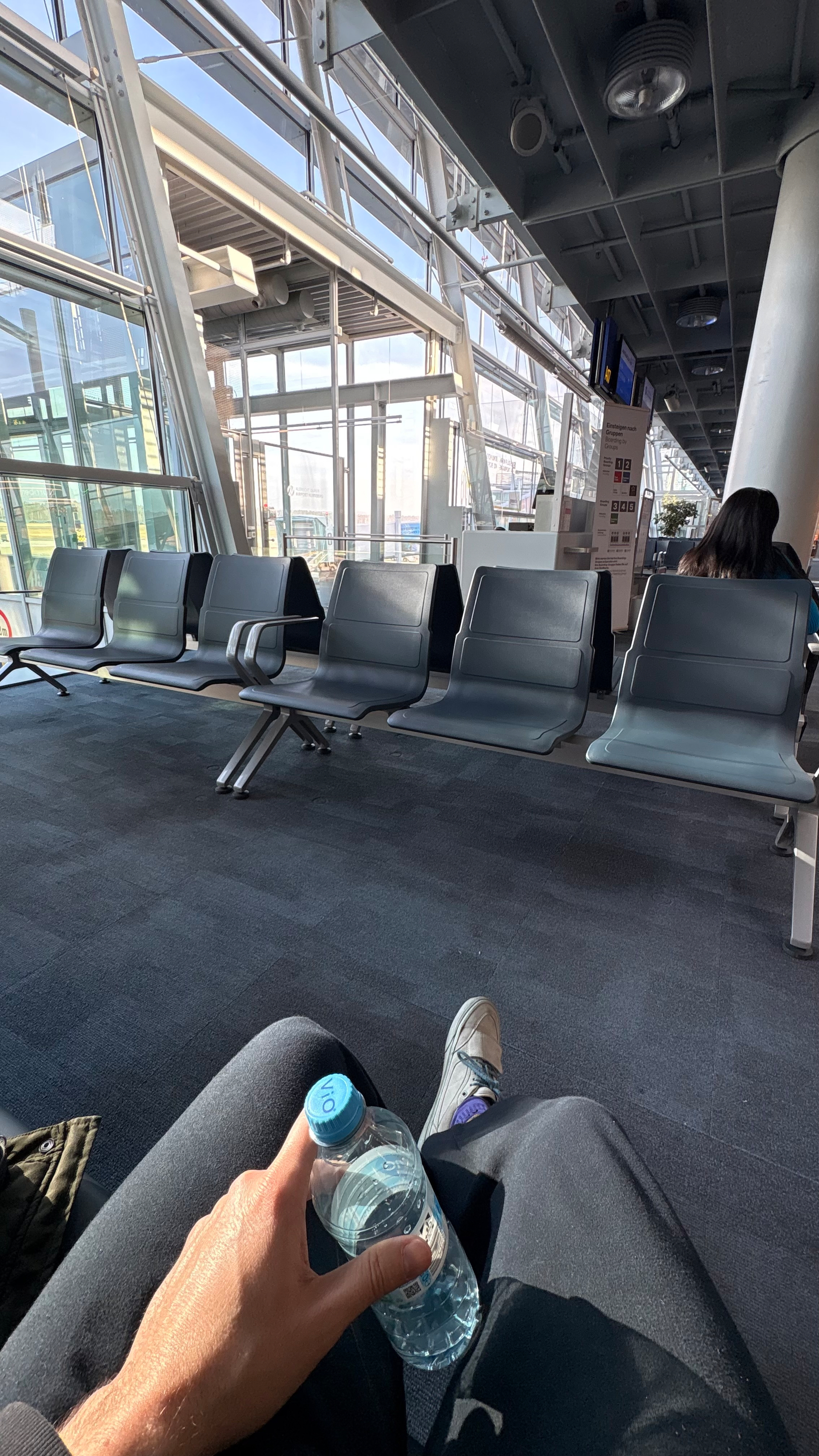 A person holding a bottle of water sits in an airport terminal with a row of unoccupied seats and large windows in the background.