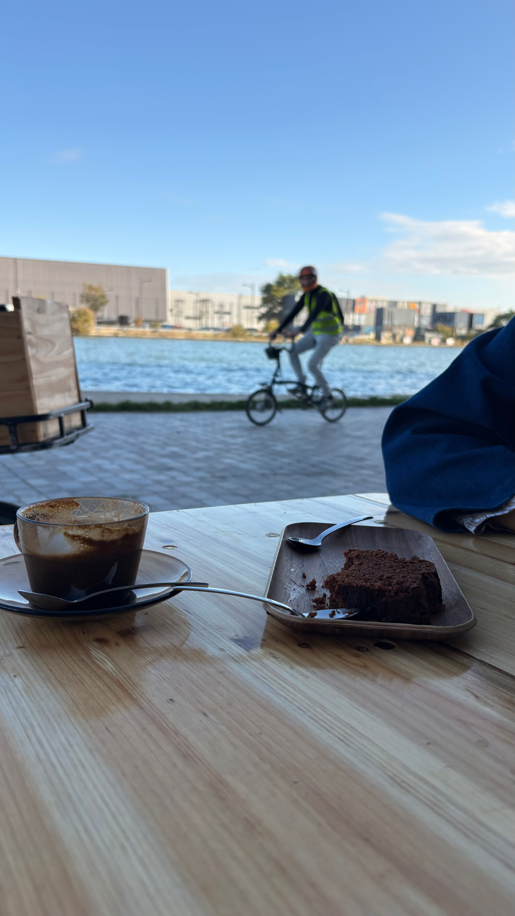 A partially eaten piece of chocolate cake and a cup of coffee rest on a wooden table with a blurred cyclist riding by a waterfront in the background.