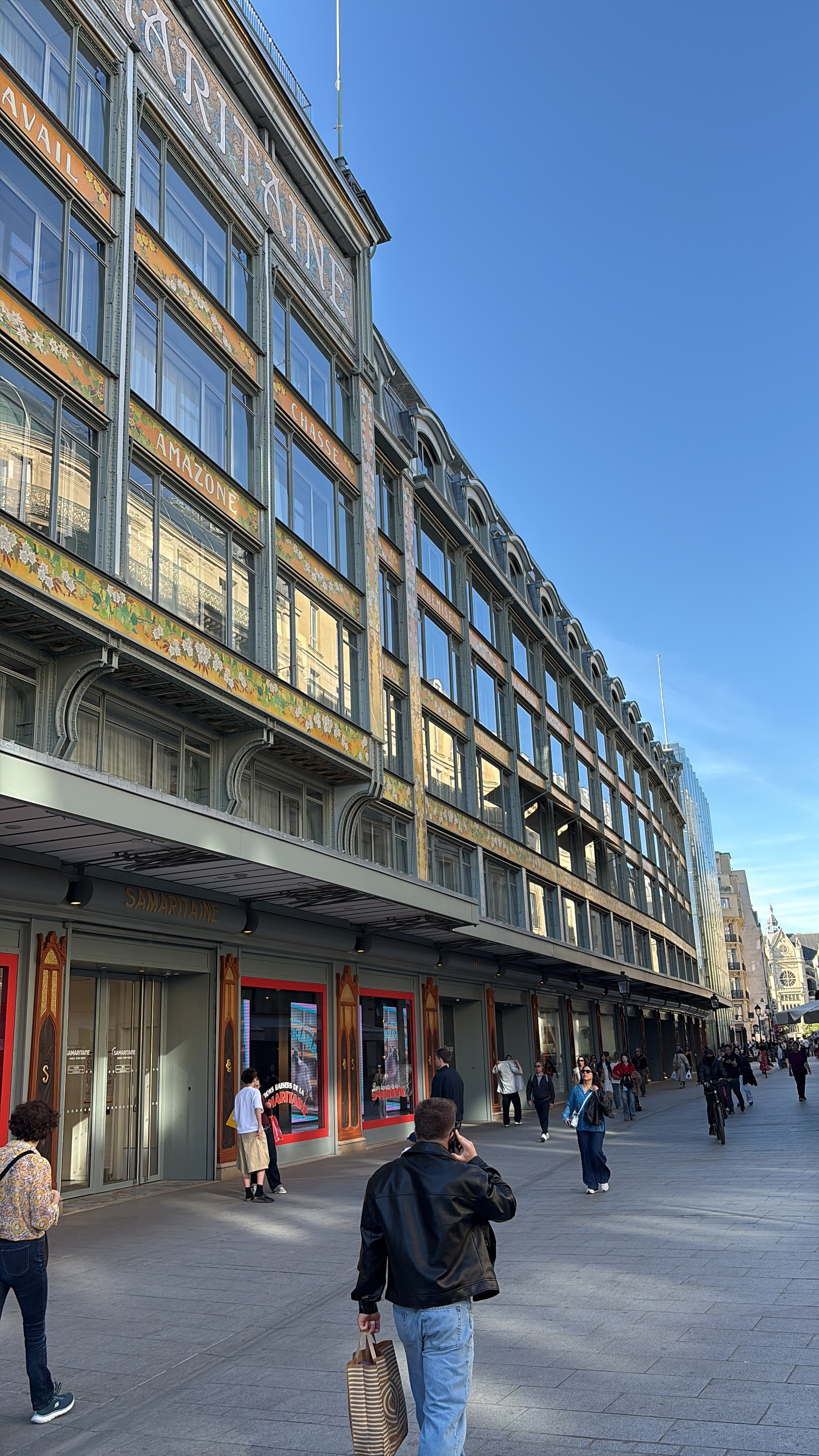 People walking along a wide street past a large, ornate building.