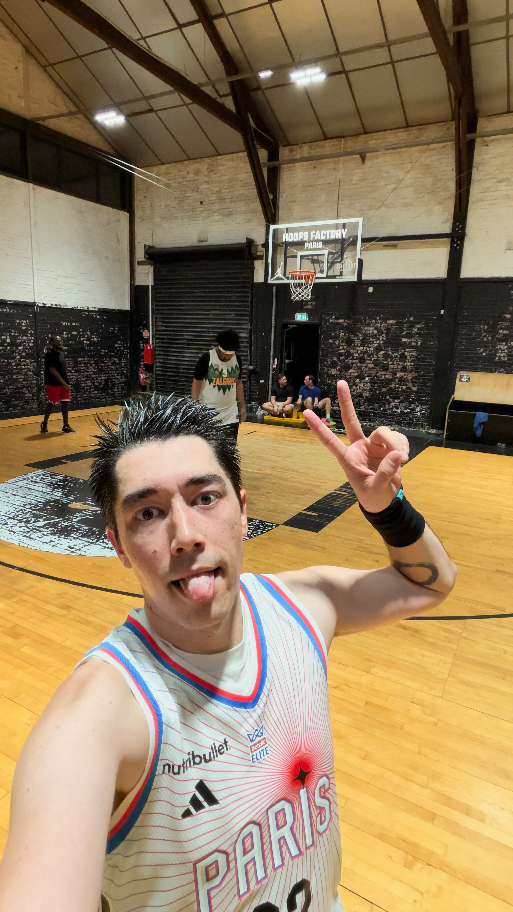 A person wearing a Paris basketball jersey is making a peace sign and sticking out their tongue on an indoor basketball court with other players in the background.