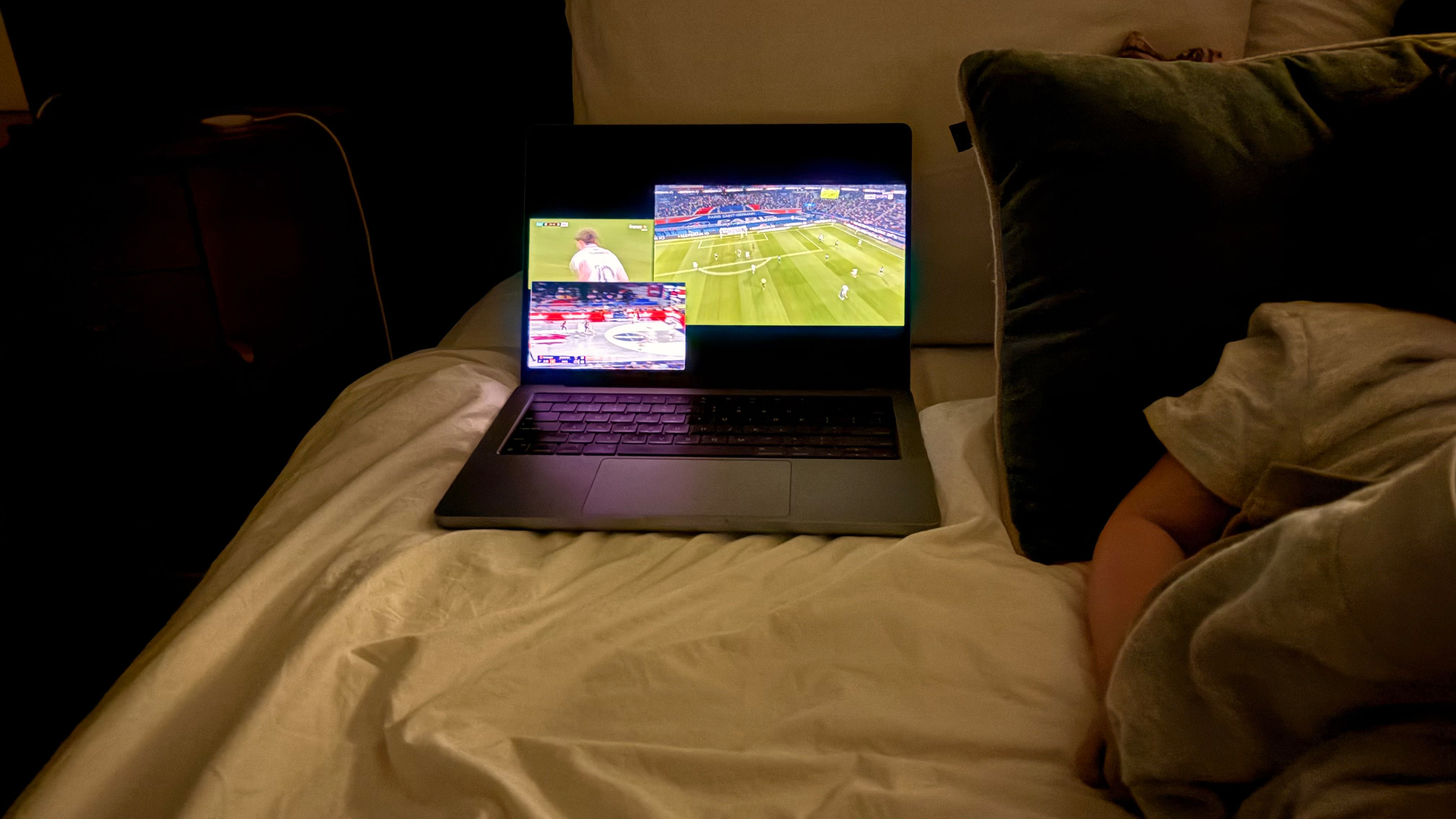 A laptop displaying multiple sports games is placed on a bed next to a child.