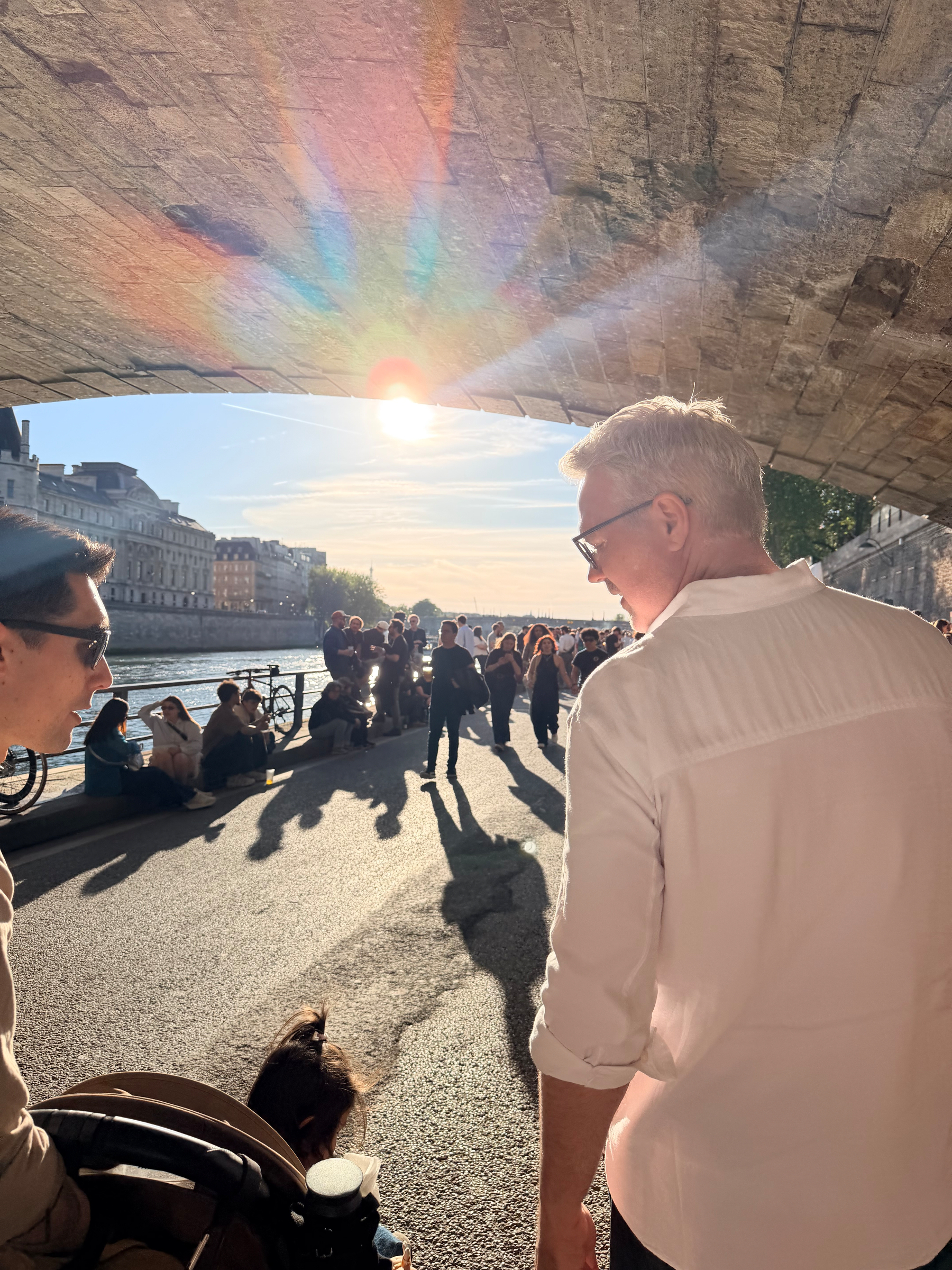 People are walking along a riverside path under a bridge with the sun shining brightly in the background.