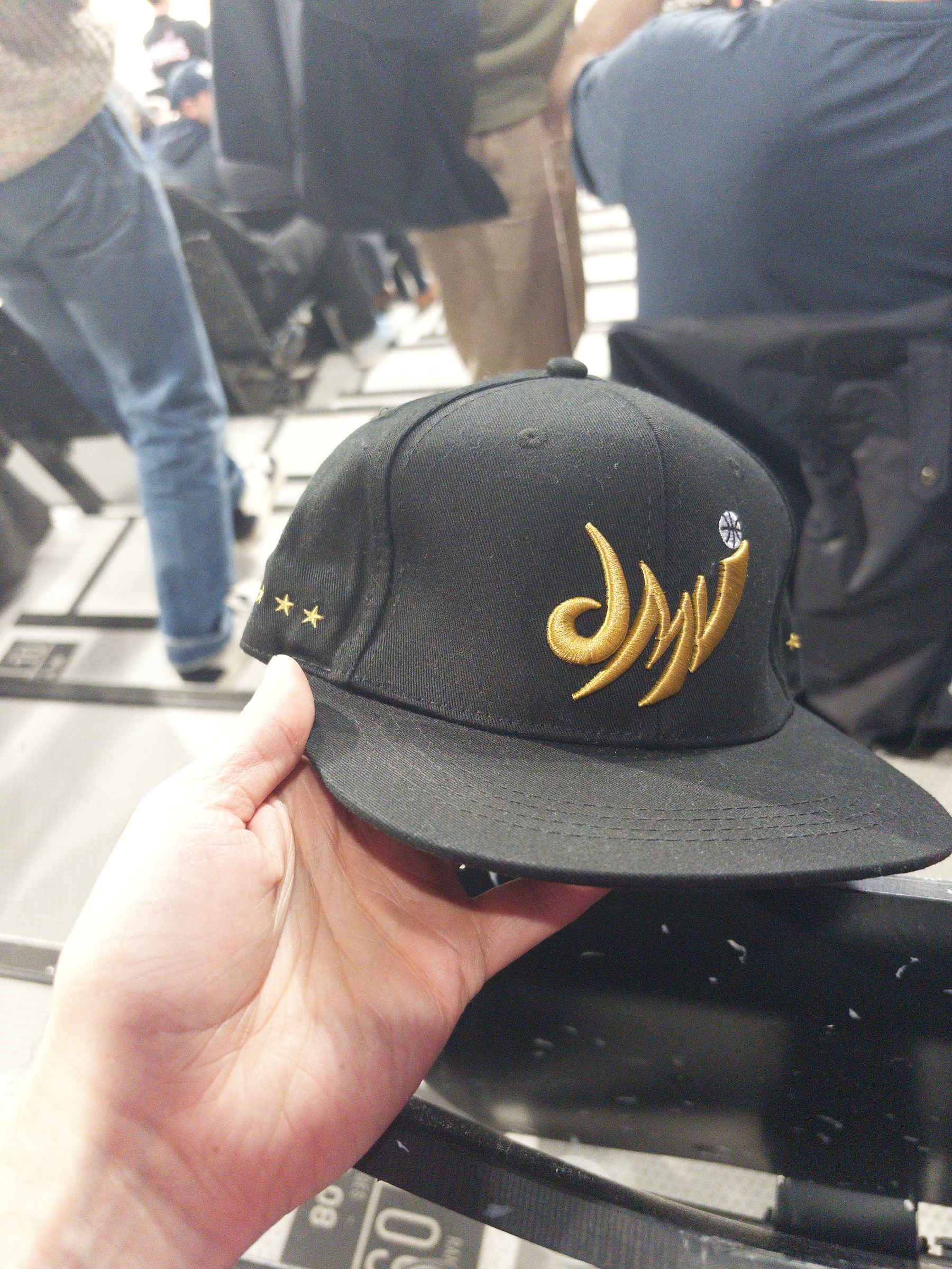 A person is holding a black baseball cap with gold embroidered designs, featuring a swirl pattern and stars, while seated among others in a stadium or arena setting.