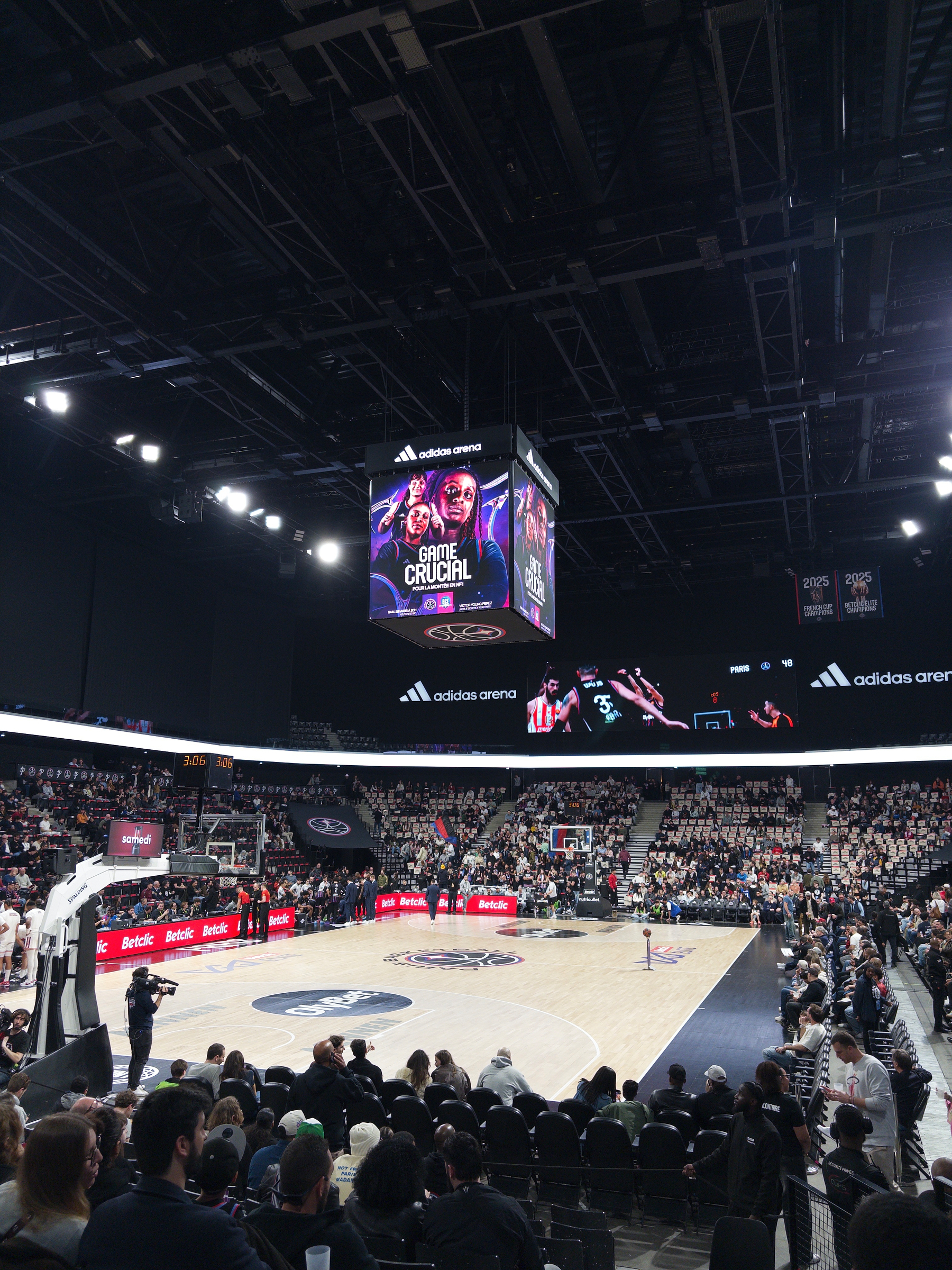 A basketball game is taking place in a crowded indoor arena with digital screens displaying the event.