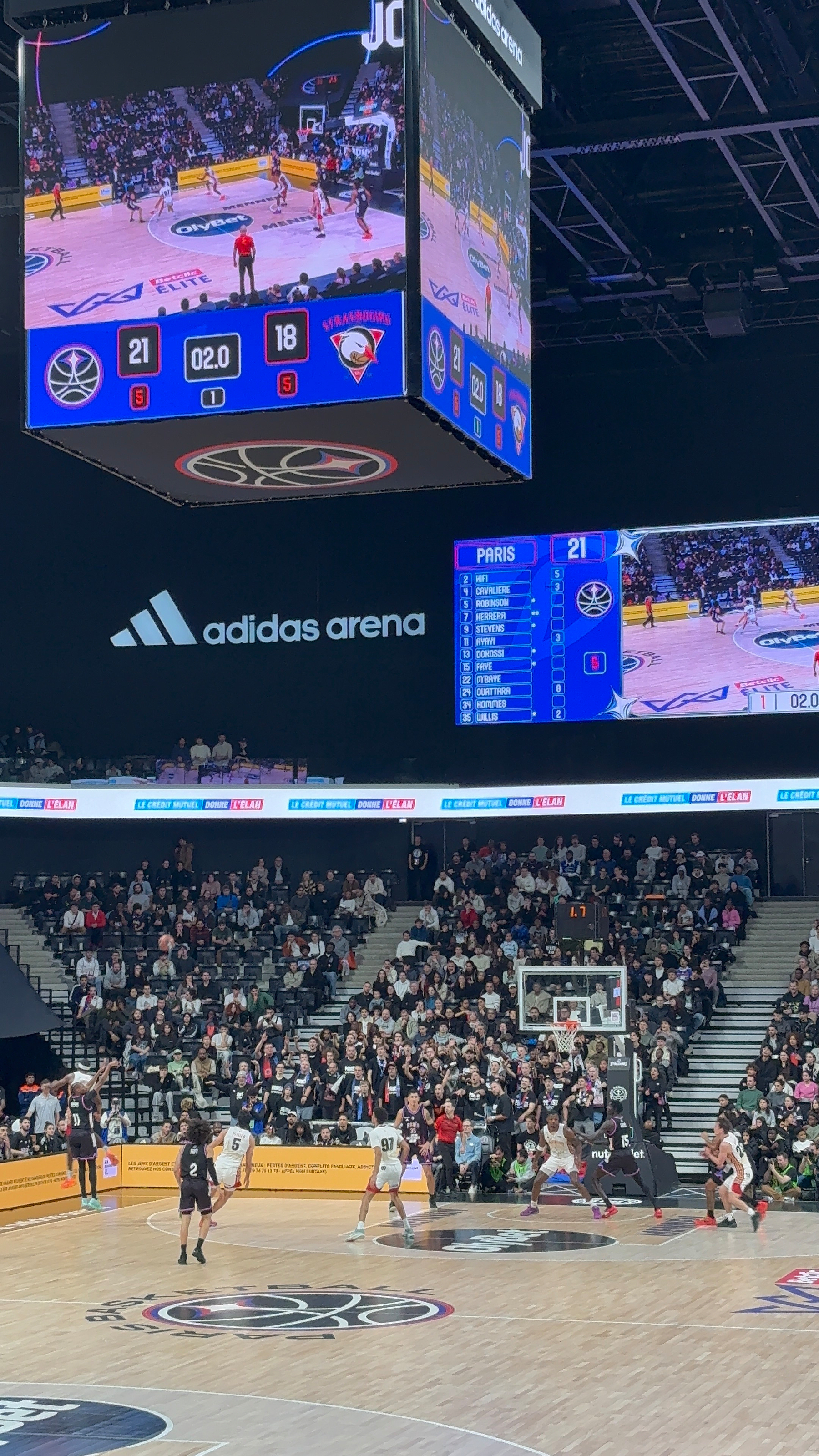 A basketball game is taking place in an arena, with a scoreboard showing a score of 21-18 and a large screen displaying the game above the court.