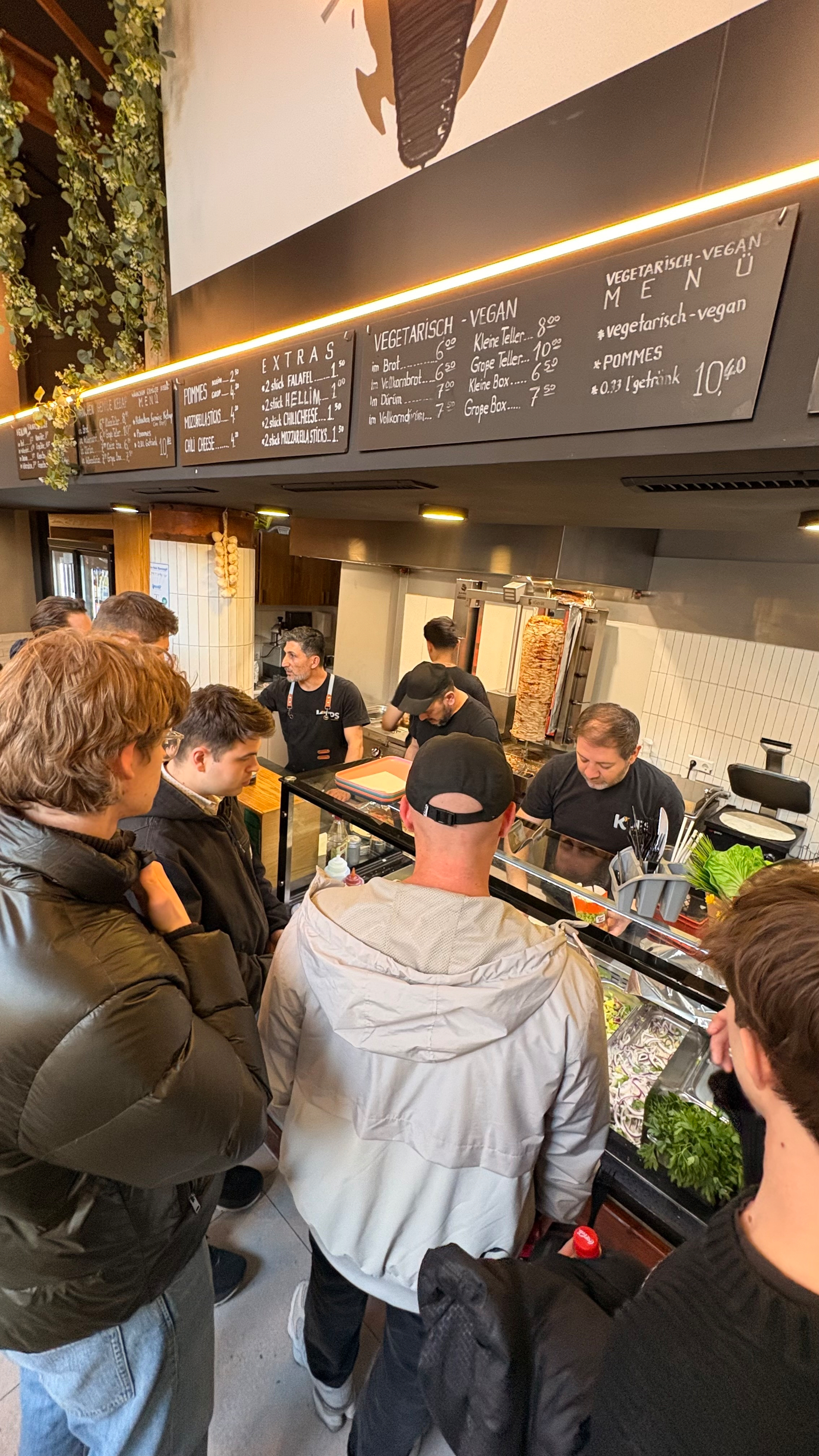 Auto-generated description: Several people are lined up at a food counter, placing orders with staff preparing meals behind the counter.