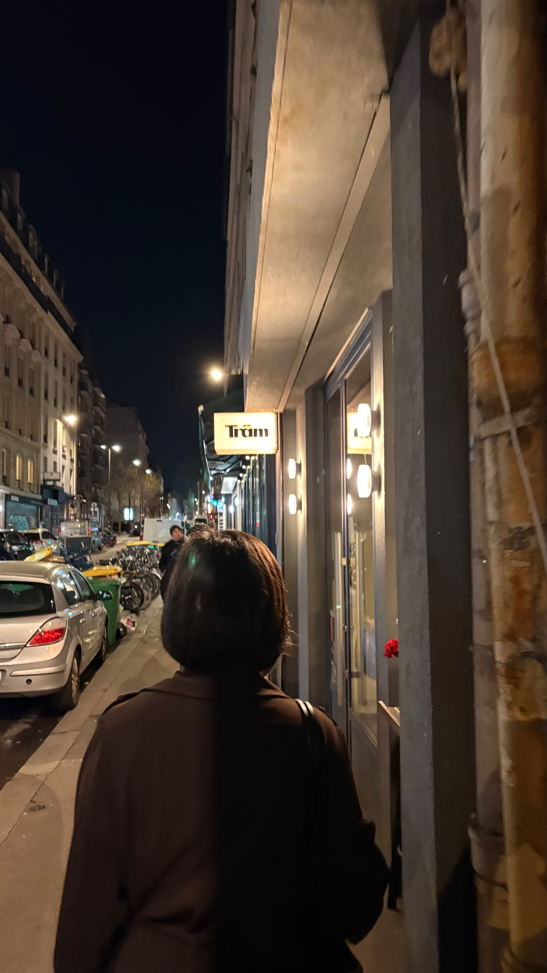 A person walks along a dimly lit street lined with parked cars and bicycles, passing by a storefront with a Trüm sign.