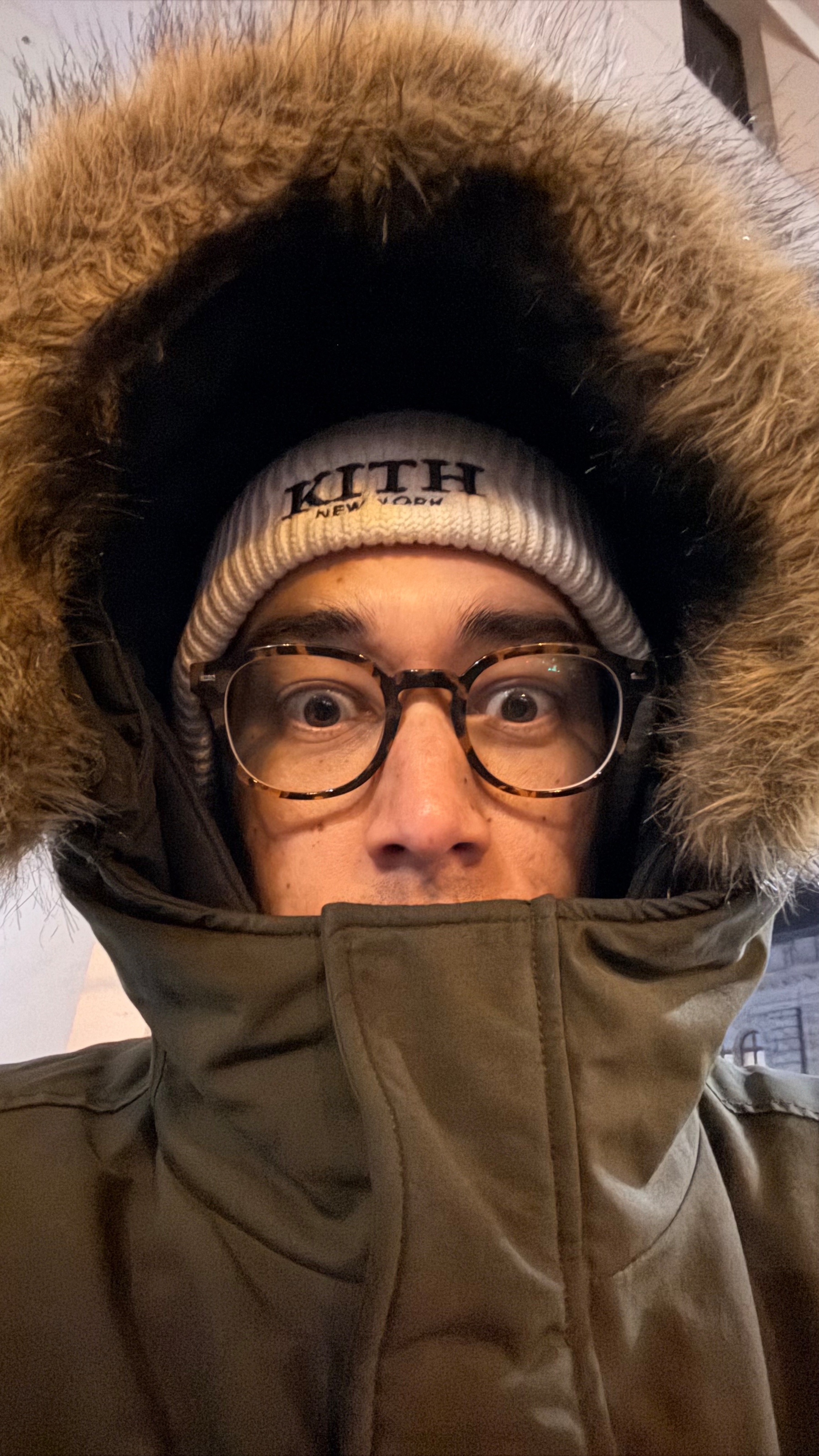 A person wearing glasses, a fur-lined hooded coat, and a beanie with wide eyes is looking at the camera.