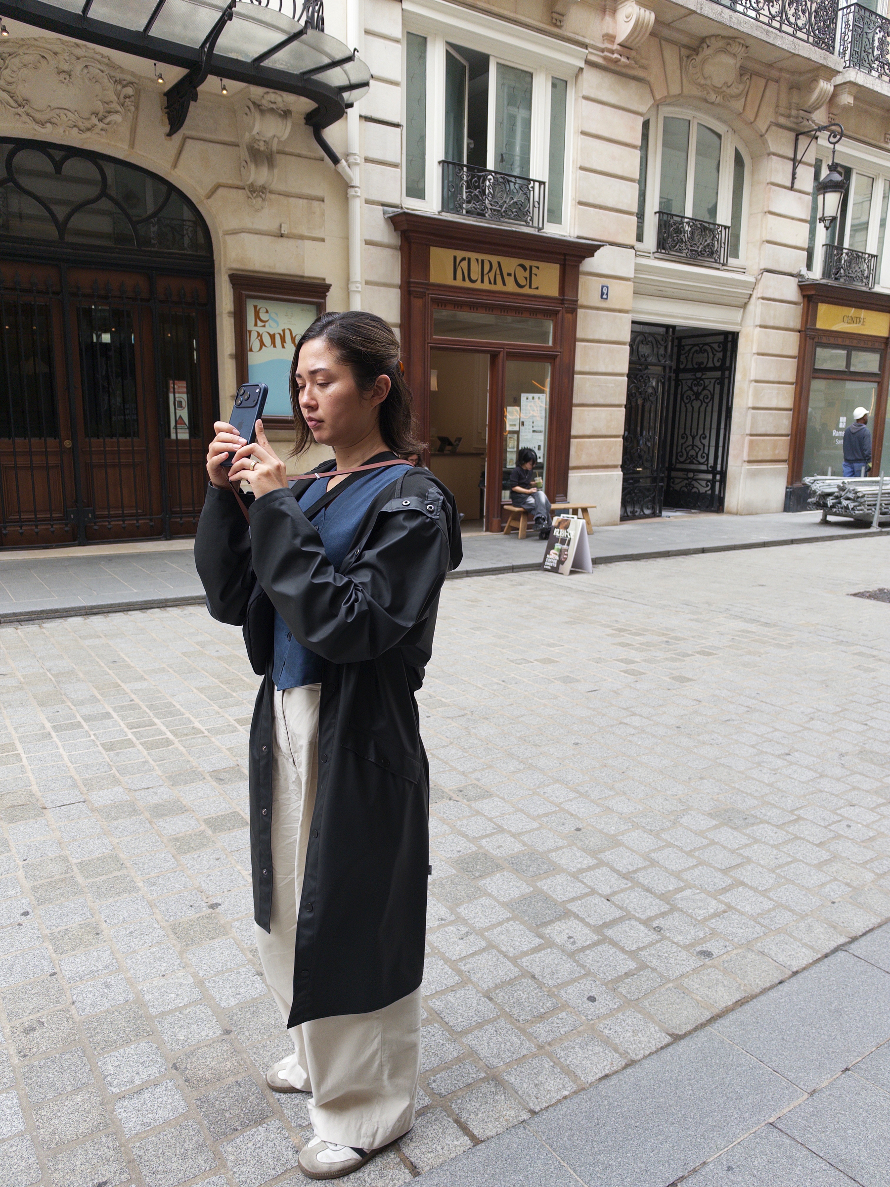 Auto-generated description: A woman dressed in a black coat and white pants is standing on a cobblestone street, using her phone to take a photo or video.