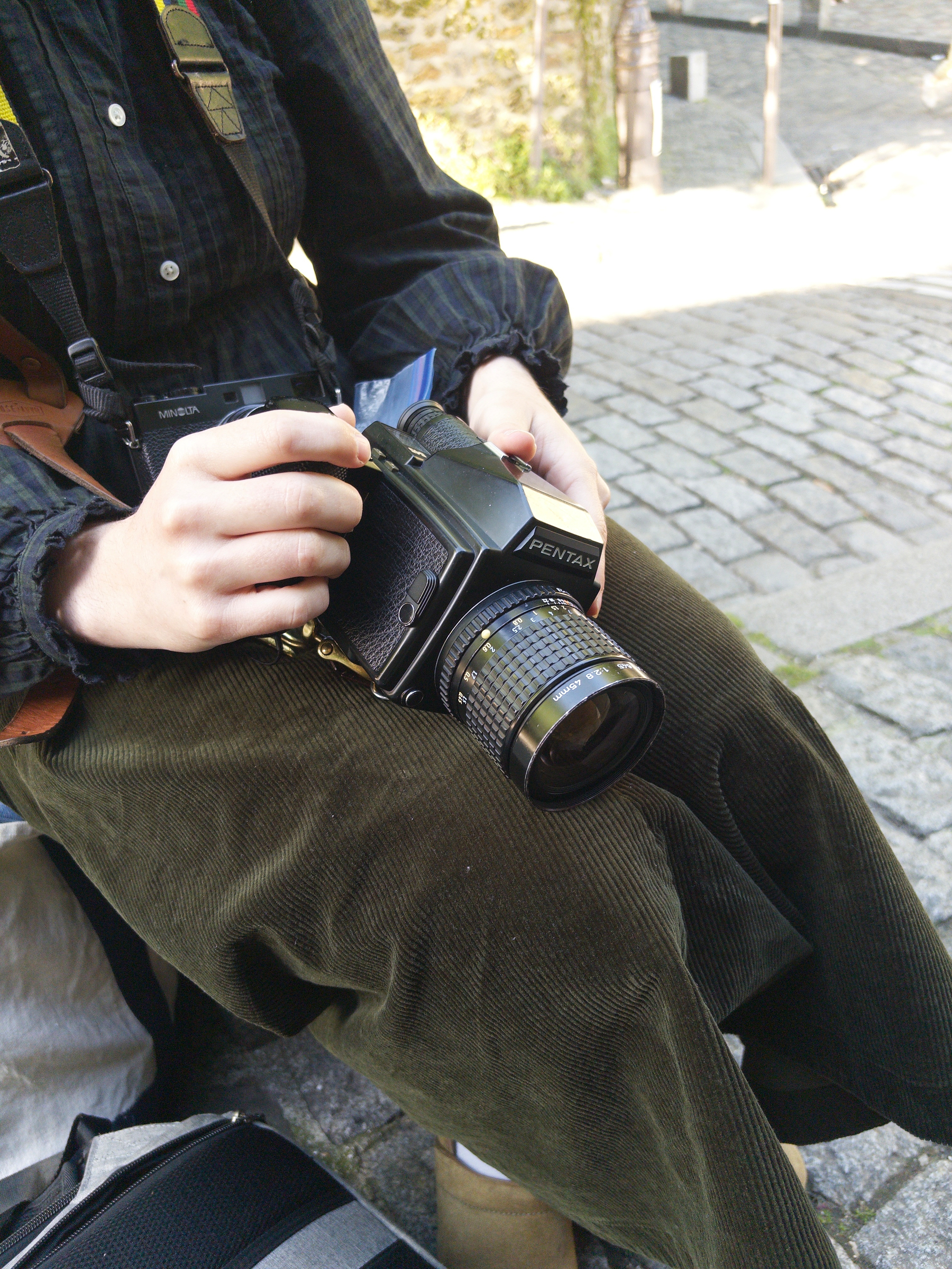 Auto-generated description: A person wearing corduroy pants and a dark shirt is sitting and holding a Pentax camera.