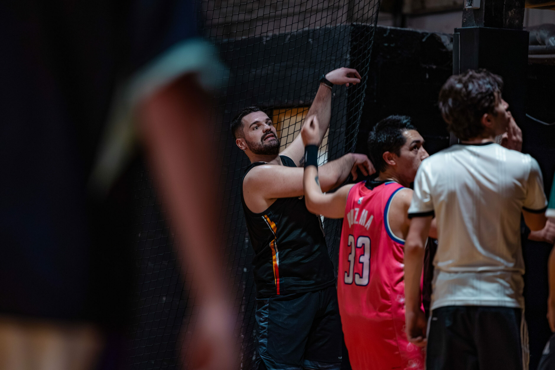 Auto-generated description: A group of men is gathered in a sports setting, with one wearing a basketball jersey and another leaning against a netted wall.