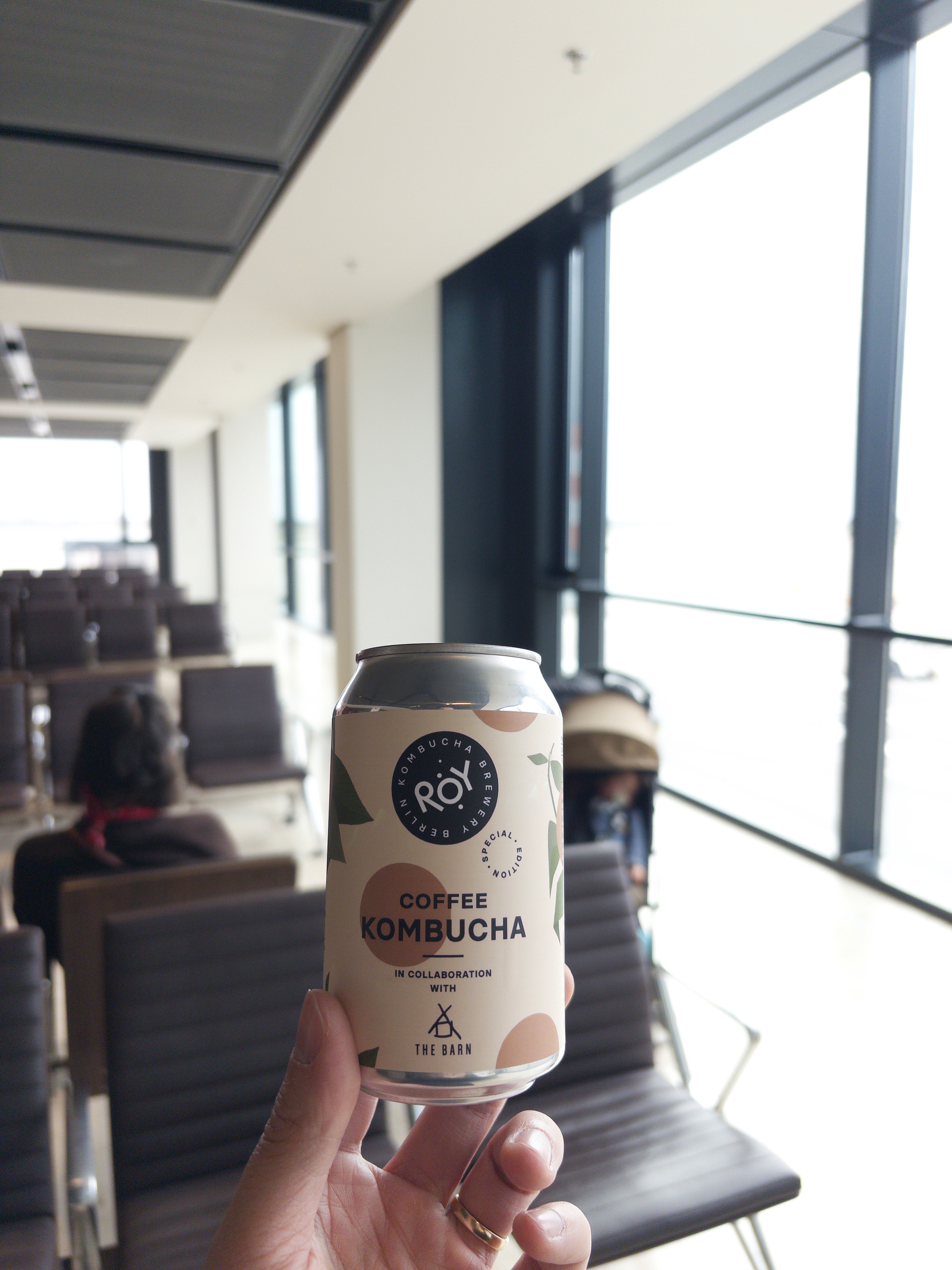 Auto-generated description: A hand holding a can of coffee kombucha in an airport seating area with several chairs and large windows.