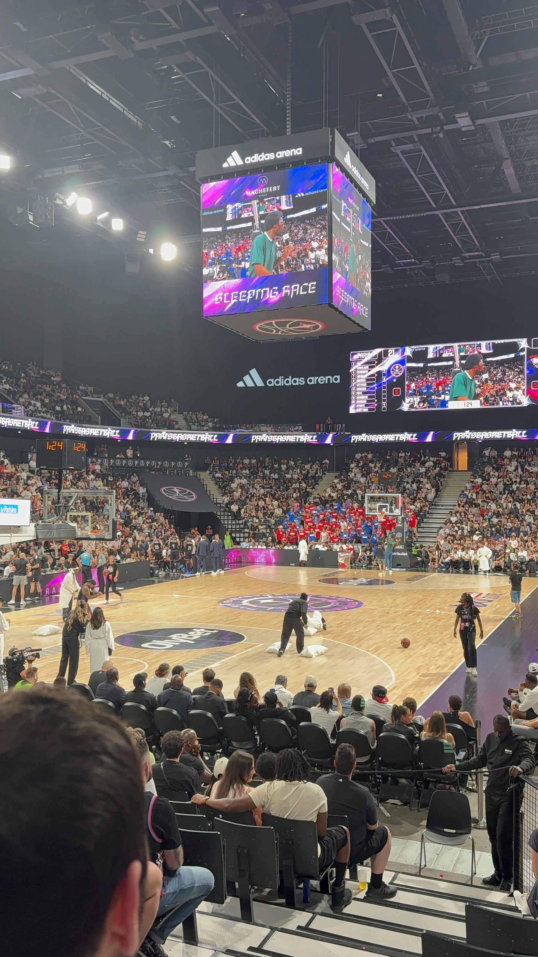 A crowded indoor arena hosts a basketball game with spectators watching from the stands and large screens displaying the event.
