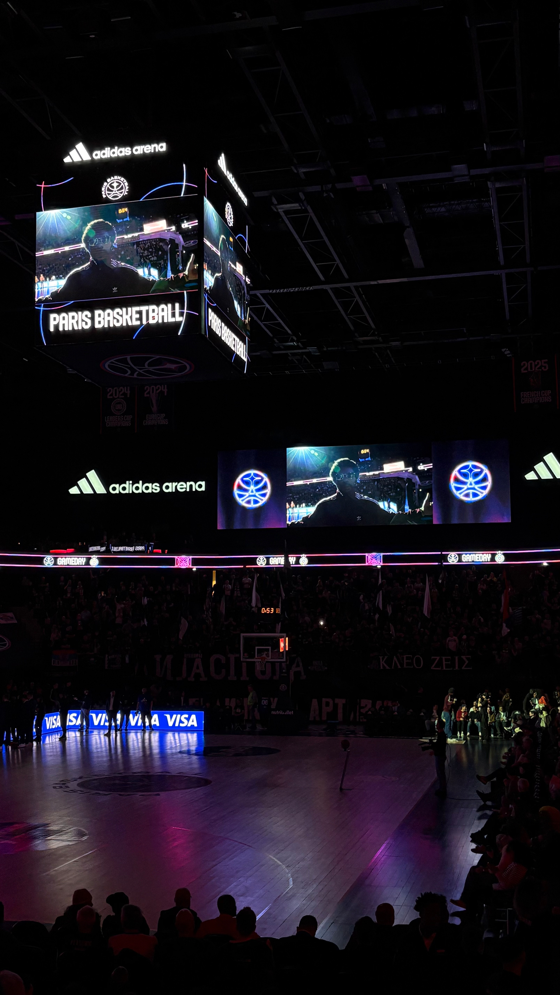 A basketball arena features a large scoreboard with Paris Basketball displayed, and a crowd watching attentively.