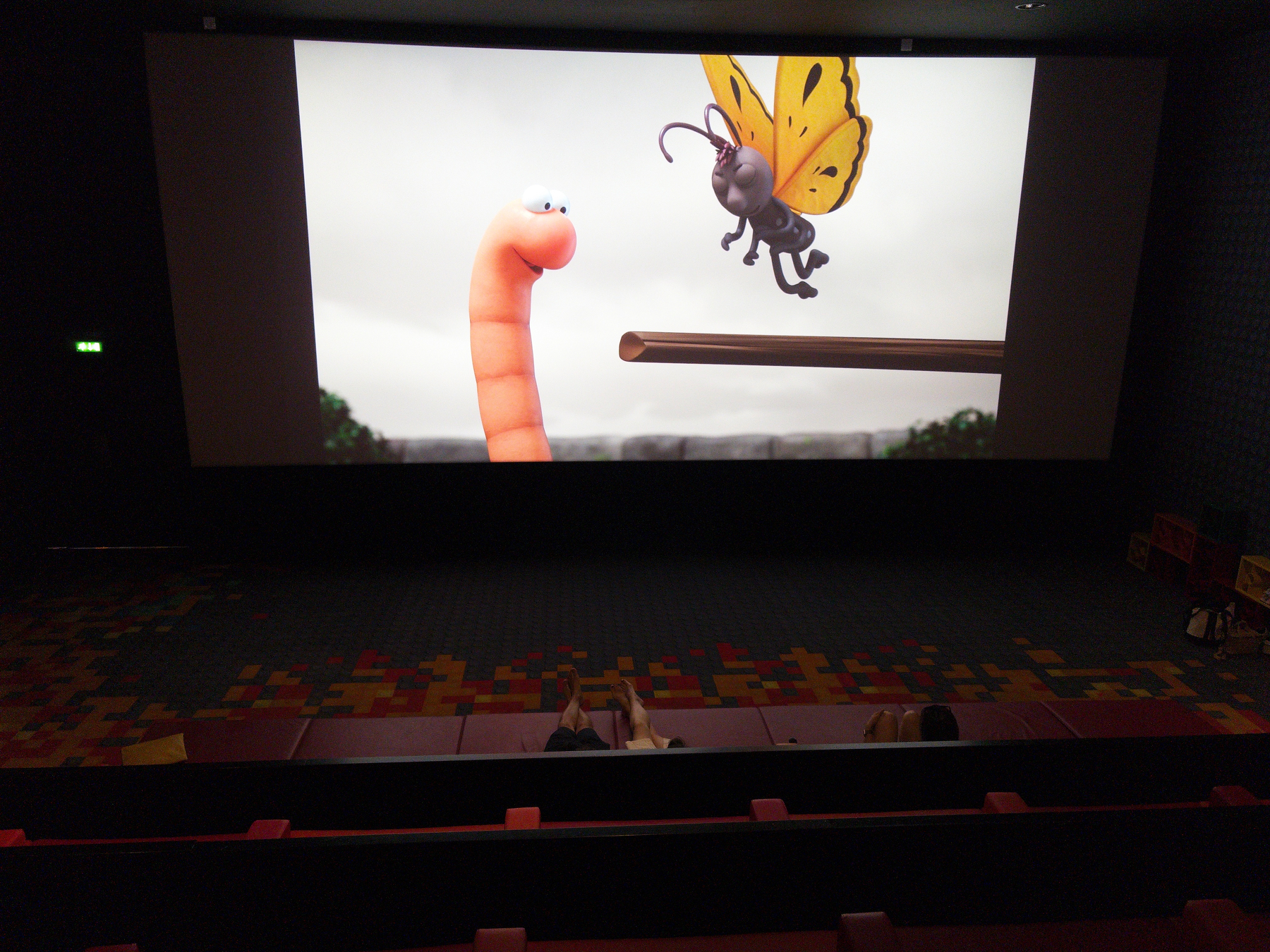 A scene from an animated movie featuring a talking worm and a butterfly is being projected in a movie theater with a few people seated in the audience.