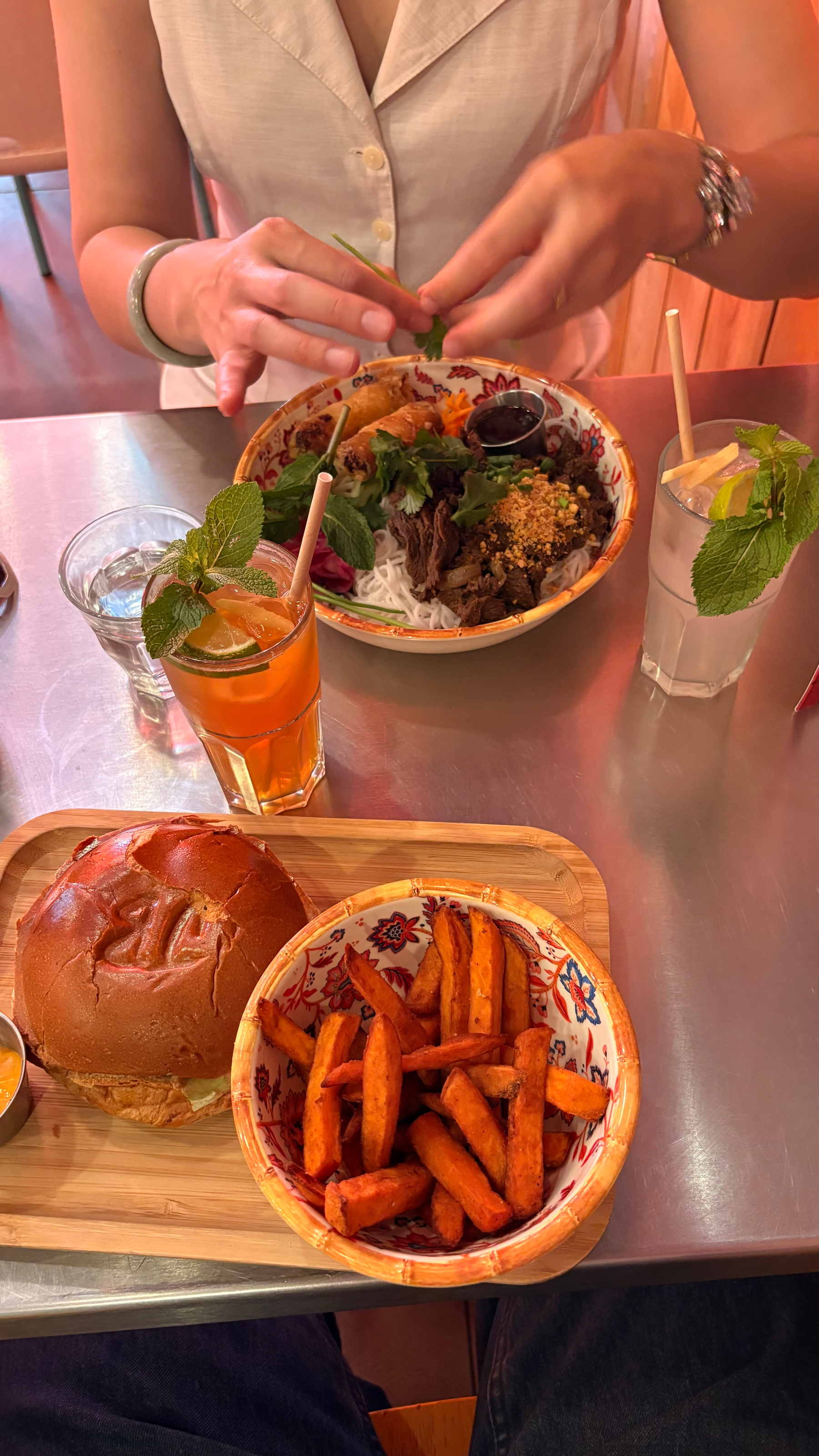 A person is seated at a table with a burger, sweet potato fries, and two drinks, while preparing a rice noodle bowl with vegetables and meat.