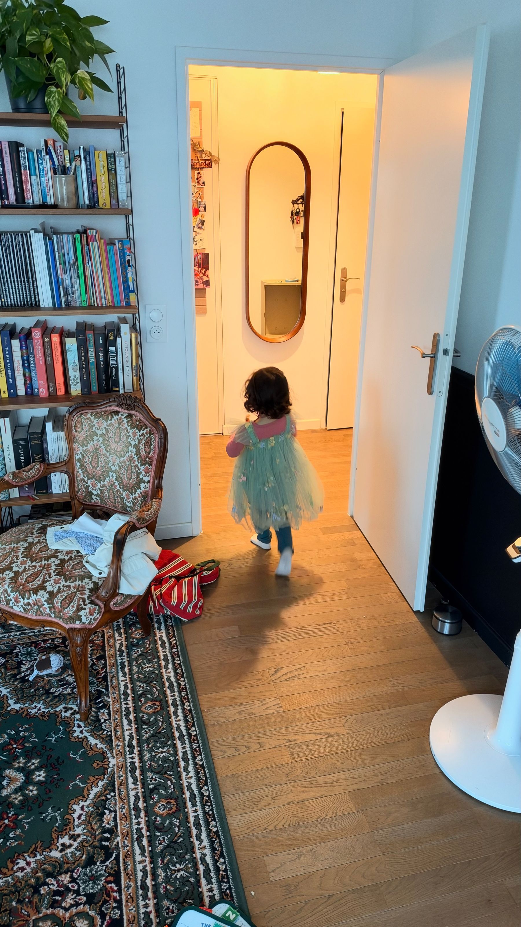 A young child wearing a green dress is walking into a brightly lit hallway from a room filled with bookshelves, an ornate chair, a standing fan, and a patterned rug.